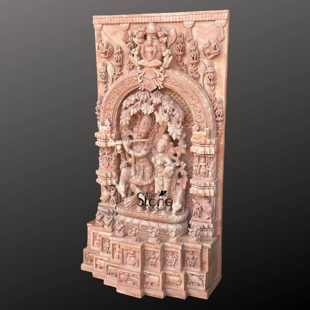Pink Stone Radha Krishna Murti for Home 4ft 5 This image features a magnificent 4-foot tall Radha Krishna Sculpture in Pink Stone with Ornate Arch Panel, showcasing exceptional craftsmanship. The entire piece is carved from beautiful natural pink stone, which imparts a warm and inviting hue. At the heart of the sculpture, Lord Krishna is depicted playing his flute under a detailed, sprawling tree, his posture graceful and serene. Standing beside him is Radha, her figure equally adorned with intricate details, her gaze directed towards Krishna, completing the divine pair. The central Radha Krishna scene is framed by a grand and highly ornate arch panel, which is deeply carved with traditional motifs, foliage, and smaller figures, creating an impression of an elaborate temple entrance or a sacred grove. Above this arch, a larger deity, possibly Vishnu, is enthroned, surrounded by additional celestial beings and intricate architectural flourishes, all meticulously carved into the pink stone. The vertical side panels flanking the central arch are densely populated with smaller sculptures, depicting various divine figures, devotees, and scenes from Hindu mythology. These detailed carvings extend downwards to the multi-tiered base, which is also embellished with a series of friezes and smaller alcoves, each holding more intricate sculptures of figures and animals. The overall effect is one of immense grandeur and spiritual depth, highlighting the beauty and narrative richness achievable in this 4-foot pink stone Radha Krishna statue, making it a luxurious and culturally significant piece.