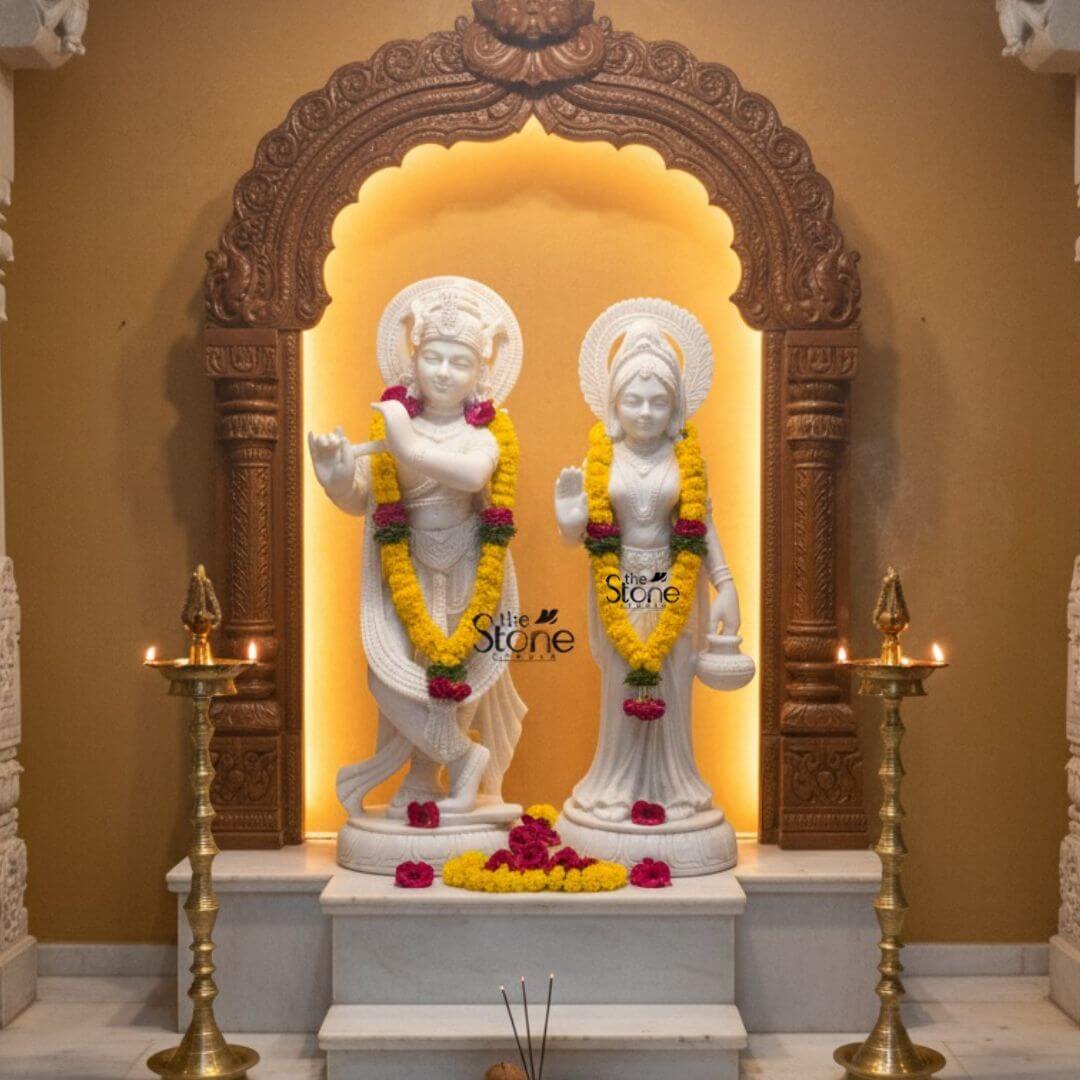 Radha Krishna Marble Stone Statue 3ft 3 This image displays a stunning Radha Krishna Temple Idol in Marble featuring a pair of 3-foot tall white marble statues of Radha and Krishna beautifully arranged on an altar in what appears to be a temple or a dedicated pooja room. The deities are presented within an intricately carved wooden archway which is illuminated from behind with a warm soft glow creating a serene and ethereal ambiance. The wooden arch itself is highly decorative featuring traditional motifs and robust pillars. On the left Lord Krishna stands in his iconic Tribhanga posture holding his flute. He is adorned with fresh garlands of yellow marigolds and red roses draped around his neck. His finely sculpted white marble form includes a detailed crown jewelry and flowing garments. A subtle circular halo is behind his head. On the right Radha stands gracefully her right hand raised in a gesture of blessing and her left hand holding a small pot. She is similarly adorned with fresh garlands of yellow marigolds and red roses, a crown, necklaces, bracelets, and a flowing sari, all intricately carved from white marble. A matching circular halo is behind her head. Both white marble statue figures stand on individual ornate lotus pedestals placed on a tiered white marble altar. In front of the statues loose yellow and red flowers are scattered and incense sticks release fragrant smoke. Two tall, elegant brass oil lamps with lit flames flank the altar adding to the sacred atmosphere. The warm golden tones of the wall and the intricate carvings create a profound and inviting setting for this revered Radha Krishna Temple Idol.