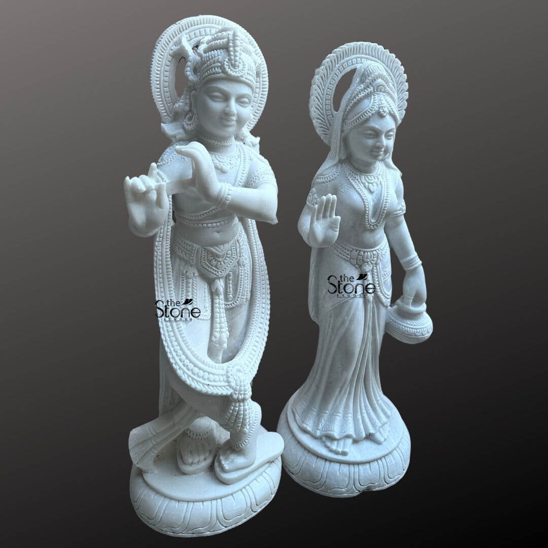 Radha Krishna Marble Stone Statue 3ft 2 This image presents a stunning Radha Krishna Temple Sculpture in Marble featuring a pair of 3-foot tall white marble statues of Radha and Krishna. Expertly carved the deities are displayed against a dark neutral background allowing the pristine white marble and intricate details to stand out making them ideal for a temple setting. On the left Lord Krishna stands in his iconic Tribhanga posture holding and playing his flute. He is richly adorned with finely sculpted jewelry, a detailed crown with a peacock feather, and flowing garments, all rendered with exceptional precision in the white marble. A long garland also encircles his form. A prominent, intricately carved circular halo (prabhavali) is behind his head, signifying his divine aura. On the right Radha stands gracefully, her right hand raised in a gesture of blessing (Abhaya Mudra), and her left hand holding a small pot or offering. She is also beautifully adorned with a crown, necklaces, bracelets, and a flowing sari, all intricately carved from the same white marble. A similar, finely carved circular halo is behind her head. Both statues stand on individual, ornate lotus pedestals, which are integrated into the overall marble carving and feature additional carved details. The smooth, polished surface of the white marble statue reflects light softly emphasizing the delicate features, the extensive carvings on their attire and jewelry, and the divine expressions of Radha and Krishna. This Radha Krishna Temple Sculpture is a beautiful embodiment of sacred love and traditional Indian artistry, perfectly suited for veneration in a temple.