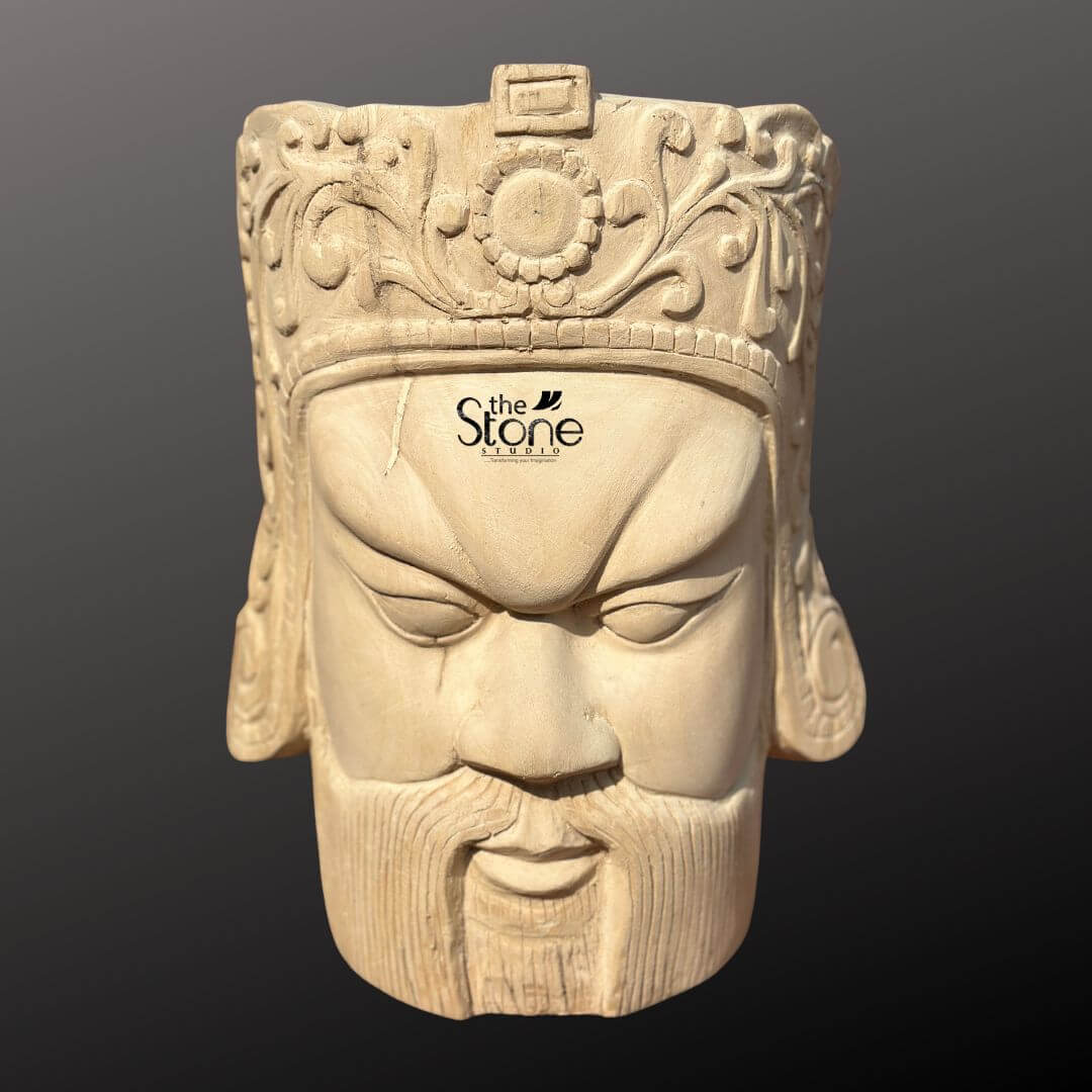 Royal Head Sculpture 12" 1 This image features a detailed 12-inch Royal Head Sculpture intricately carved from light-colored wood, showcasing a regal and stern male face. The sculpture, presented against a dark, neutral background, emphasizes the fine craftsmanship and the texture of the wood. The face itself has strong, defined features, including a prominent brow, deeply set eyes with sharp angles, a straight nose, and a closed mouth with a full, sculpted beard and mustache. The expression conveys authority and a somewhat fierce determination, fitting for a royal head. The most striking feature is the elaborate crown or headdress worn by the figure. It is adorned with detailed floral and scrollwork carvings, a central circular motif (possibly a sun or flower), and patterned bands, all carved from the same piece of wood. The carving style suggests an East Asian or ancient warrior aesthetic, with intricate patterns on the sides of the headpiece. The natural grain of the wood is visible throughout the sculpture, particularly on the face and beard, adding to its authentic and rustic yet refined appearance. The wooden head sculpture truly stands out as a unique piece of art, blending regal power with natural material.