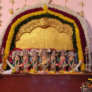 This image displays a vibrant Sati Aasra Devi Murti Set arranged on an altar, featuring a row of seven identically styled female deities, likely carved from black granite stone and then vividly colored. These figures are set within a golden arched frame, which is itself magnificently decorated with multiple garlands of fresh flowers in white, yellow, and red, creating a festive and devotional atmosphere. The flower garlands form a vibrant rainbow arch above the deities, extending down the sides. Each Devi figure is adorned with traditional, stylized features: round faces, large, dark eyes, and red bindis on their foreheads. They all wear elaborate crowns, necklaces, and earrings, all painted to mimic gold and precious stones. Their attire alternates between rich red and deep green traditional Indian garments, each with subtle patterns. They are depicted sitting or standing, with some holding a golden trident (trishul) and others having their hands clasped in prayer or blessing. The Devi figures are placed on a wooden altar or shelf, which is decorated with fresh offerings including fruits (such as apples and pomegranates), loose yellow flowers, and possibly other puja items. Small traditional oil lamps (diyas) flank the set on either side, their soft glow adding to the sacred ambiance. The entire arrangement, with its rich colors, intricate details, and fresh floral decorations, clearly represents a revered Sati Aasra Devi Murti Set designed for worship, embodying tradition and spiritual beauty.