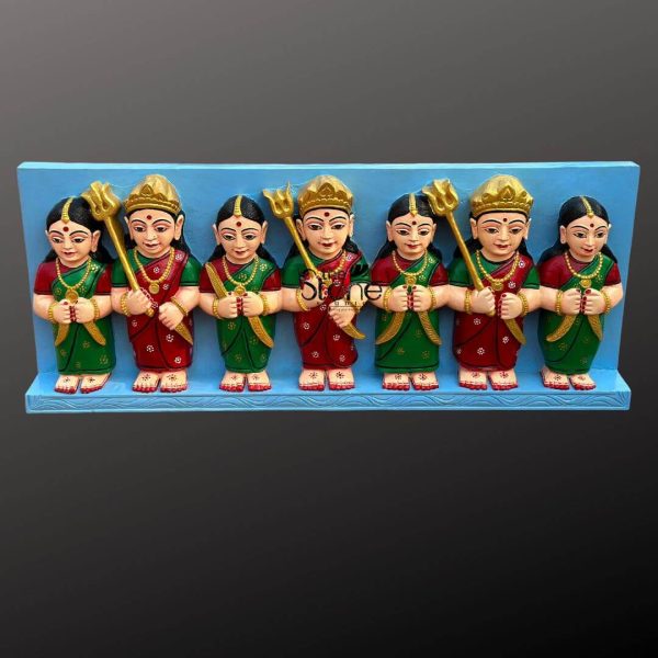 This image displays a vibrant Sati Aasra Devi Statue panel, depicting seven identical female deities, likely carved from black granite stone and then brightly colored, measuring approximately 4 feet in length. Each of the seven Devi figures stands side-by-side, holding hands, creating a sense of unity and strength. The figures are stylized, with round faces, large expressive eyes, and red bindis on their foreheads. They are adorned with golden crowns, matching golden jewelry around their necks, and small earrings. Their attire alternates between red and green traditional Indian garments, each featuring delicate patterns, possibly representing saris or ghagras. Their small feet are visible at the base, decorated with white floral motifs. The two central figures and the two figures second from each end each hold a golden trident (trishul) in their outer hand, signifying divine power and protection. The figures on the far left and far right have both hands clasped together, suggesting a gesture of prayer or reverence. The entire ensemble is mounted on a light blue rectangular panel, which serves as a backdrop and a base, making the colorful statues pop. The black granite stone (coloured) gives them a robust base while the painted details bring them to life, highlighting their significance as a Sati Aasra Devi worship art piece.