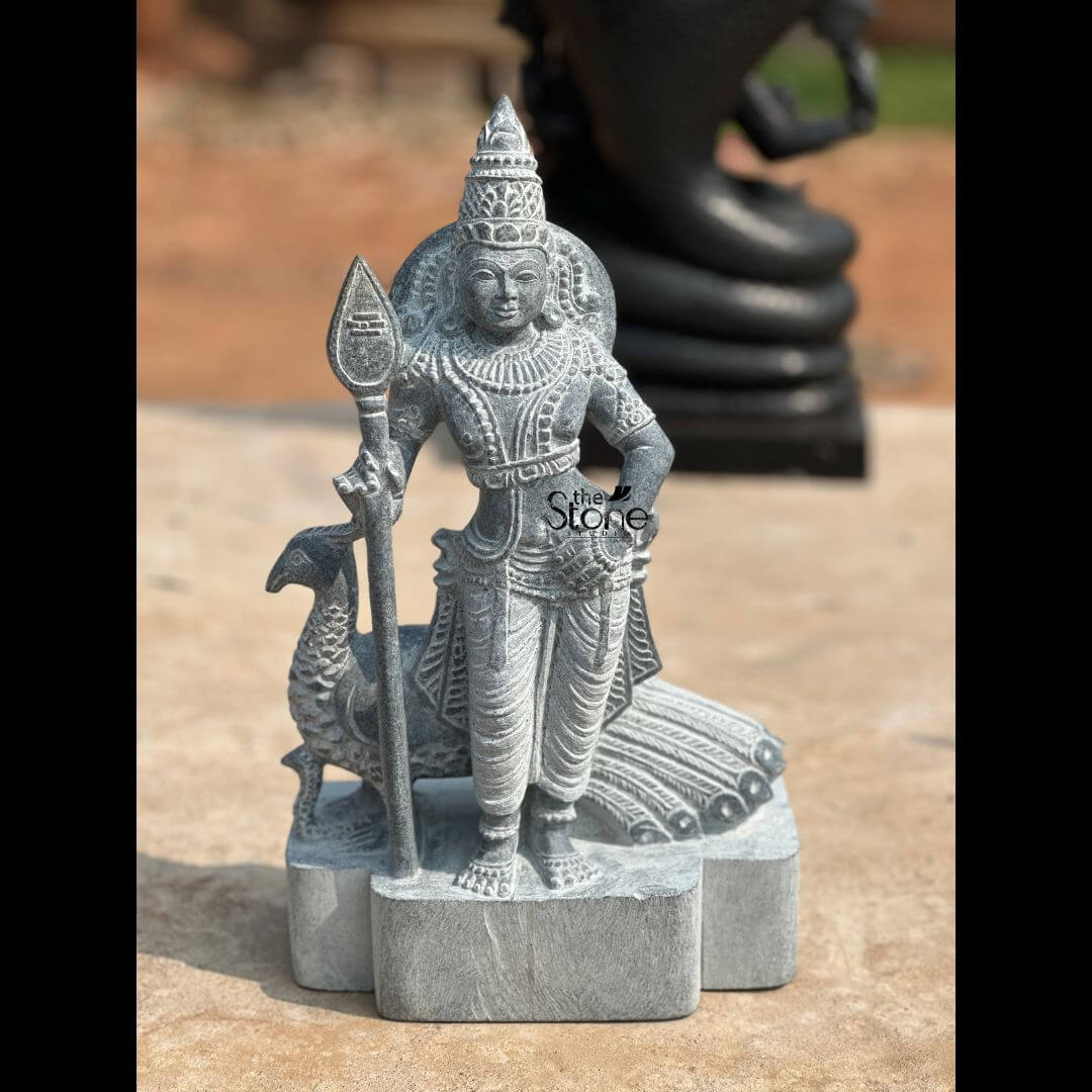 Lord Skanda Murugan Stone Statue 15" 3 An expertly hand-carved 15-inch Skanda Murugan idol, masterfully sculpted from premium natural blackstone with a rustic, weathered finish. The deity is depicted in a majestic standing posture, holding his iconic sacred spear, the Vel, in his right hand, which symbolizes divine wisdom and the destruction of negative forces. Lord Murugan is beautifully adorned with a towering tiered crown (mukut), layered necklaces, armlets, and traditional garments with realistically carved fabric folds. Standing faithfully behind the deity is his divine vehicle, the peacock, with its head visible on the left and its long tail feathers delicately rendered in relief on the right. The entire sculpture rests on a multi-lobed stone pedestal, making it a perfect spiritual centerpiece for a home mandir, garden landscape, or meditation sanctuary. The deep charcoal tones and grainy texture of the blackstone statue reflect timeless Indian craftsmanship, designed to invite strength, victory, and protection into any sacred or living space.