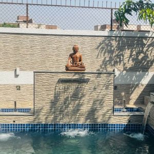 This image features a serene 2-foot Stone Buddha Statue specifically a brown sandstone Buddha beautifully integrated into a modern outdoor water feature likely in Lonavala Maharashtra. The Buddha statue is seated in a meditative posture, with legs crossed and hands resting in his lap atop a textured wall. The statue, carved from reddish-brown sandstone, has a classic smooth finish depicting the Buddha's serene face with downcast eyes, coiled hair (ushnisha), and elongated earlobes. The texture and color of the sandstone blend naturally with the surroundings. The Buddha statue is positioned centrally on a prominent, textured cream-colored stone wall, which serves as a backdrop for a cascading waterfall feature. Water gently pours from a horizontal spout in the middle of this wall, creating a soothing curtain of water that falls into a sparkling blue-tiled swimming pool or pond below. To the right, another smaller waterfall feature is visible, also flowing into the pool. The pool itself is lined with blue and white tiles, adding a refreshing visual element. Above the main Buddha wall, a lighter cream-colored textured wall extends, topped by a metal chain-link fence, suggesting a boundary. The scene is bathed in natural sunlight, with dappled shadows from an unseen tree falling across the textured walls, enhancing the peaceful and contemplative atmosphere of this outdoor installation. This Brown sandstone Buddha statue creates a tranquil focal point for relaxation and spiritual reflection.