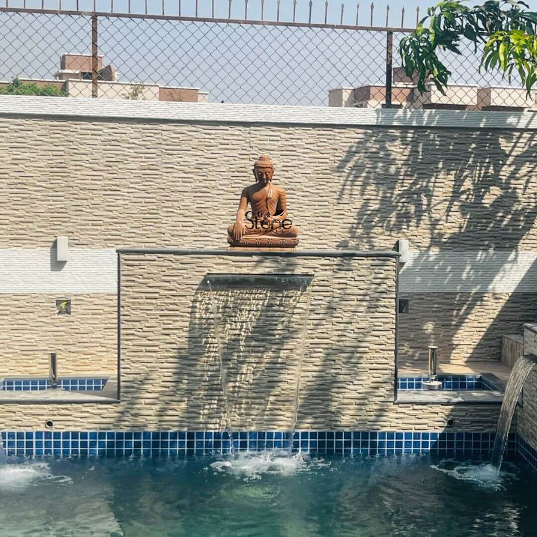 Gautam Buddha Small Statue 2ft 6 This image features a serene 2-foot Stone Buddha Statue specifically a brown sandstone Buddha beautifully integrated into a modern outdoor water feature likely in Lonavala Maharashtra. The Buddha statue is seated in a meditative posture, with legs crossed and hands resting in his lap atop a textured wall. The statue, carved from reddish-brown sandstone, has a classic smooth finish depicting the Buddha's serene face with downcast eyes, coiled hair (ushnisha), and elongated earlobes. The texture and color of the sandstone blend naturally with the surroundings. The Buddha statue is positioned centrally on a prominent, textured cream-colored stone wall, which serves as a backdrop for a cascading waterfall feature. Water gently pours from a horizontal spout in the middle of this wall, creating a soothing curtain of water that falls into a sparkling blue-tiled swimming pool or pond below. To the right, another smaller waterfall feature is visible, also flowing into the pool. The pool itself is lined with blue and white tiles, adding a refreshing visual element. Above the main Buddha wall, a lighter cream-colored textured wall extends, topped by a metal chain-link fence, suggesting a boundary. The scene is bathed in natural sunlight, with dappled shadows from an unseen tree falling across the textured walls, enhancing the peaceful and contemplative atmosphere of this outdoor installation. This Brown sandstone Buddha statue creates a tranquil focal point for relaxation and spiritual reflection.
