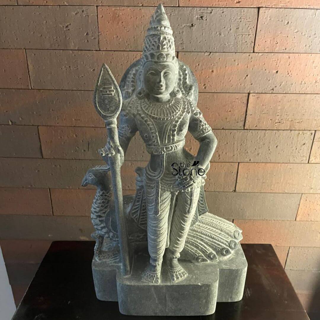 Lord Skanda Murugan Stone Statue 15" 7 An exquisitely hand-carved 15-inch Lord Skanda blackstone statue serves as a serene spiritual centerpiece, representing the rich tradition of stone statues in Kerala. The deity, also known as Lord Murugan or Kartikeya, is depicted in a majestic standing posture, holding his iconic sacred spear (Vel) in his right hand, which symbolizes divine wisdom and protection. The sculpture is masterfully crafted from premium natural black stone, featuring a rustic, grey finish that highlights the intricate textures of the carving. Lord Skanda is adorned with a towering tiered crown (mukut), layered necklaces, and traditional ornaments. His divine vehicle, the peacock, is delicately carved behind him, with its feathers fanning out in a graceful relief pattern. The idol is elegantly positioned on a dark wooden surface against a backdrop of a warm, reddish-brown brick wall. The side lighting emphasizes the three-dimensional depth of the stone work, showcasing the fine detail in the deity’s facial features and the realistic folds of his traditional attire. This compact blackstone Murugan idol perfectly illustrates the blend of ancient temple craftsmanship and modern home decor, inviting an atmosphere of strength, peace, and auspicious energy into a Kerala household sanctuary.
