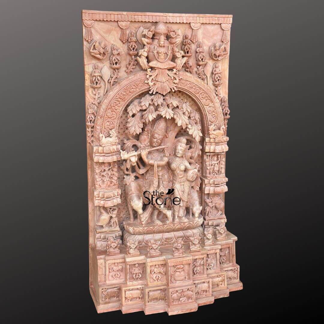 Pink Stone Radha Krishna Murti for Home 4ft 7 This image features a magnificent 4-foot tall Radha Krishna Sculpture in Pink Stone with Ornate Arch Panel, showcasing exceptional craftsmanship. The entire piece is carved from beautiful natural pink stone, which imparts a warm and inviting hue. At the heart of the sculpture, Lord Krishna is depicted playing his flute under a detailed, sprawling tree, his posture graceful and serene. Standing beside him is Radha, her figure equally adorned with intricate details, her gaze directed towards Krishna, completing the divine pair. The central Radha Krishna scene is framed by a grand and highly ornate arch panel, which is deeply carved with traditional motifs, foliage, and smaller figures, creating an impression of an elaborate temple entrance or a sacred grove. Above this arch, a larger deity, possibly Vishnu, is enthroned, surrounded by additional celestial beings and intricate architectural flourishes, all meticulously carved into the pink stone. The vertical side panels flanking the central arch are densely populated with smaller sculptures, depicting various divine figures, devotees, and scenes from Hindu mythology. These detailed carvings extend downwards to the multi-tiered base, which is also embellished with a series of friezes and smaller alcoves, each holding more intricate sculptures of figures and animals. The overall effect is one of immense grandeur and spiritual depth, highlighting the beauty and narrative richness achievable in this 4-foot pink stone Radha Krishna statue, making it a luxurious and culturally significant piece.