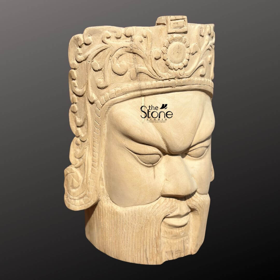 Royal Head Sculpture 12" 6 This image features a detailed 12-inch Traditional King Face Statue, meticulously carved from light-colored wood. The sculpture depicts a regal male head with a commanding and somewhat fierce expression, presented against a dark, neutral background, emphasizing the fine craftsmanship and the natural texture of the wood. The face has strong, defined features, consistent with a Traditional King or warrior aesthetic: a prominent brow, deeply set, narrow eyes that gaze downward, a straight nose, and a firmly closed mouth with a full, sculpted beard and mustache. The expression conveys authority, strength, and a sense of ancient power. Crowning the head is an elaborate headdress or crown, richly detailed with intricate floral patterns, swirling motifs, and a central circular emblem, possibly a sun or a stylized flower. The precision of the carving is evident in every element, all meticulously shaped from the single piece of wood. The natural grain of the wood is visible throughout the sculpture, particularly on the face and beard, adding to its authentic and rustic yet refined appearance. This wooden head sculpture serves as an impactful and artistic focal point, embodying historical regality and masterful carving.