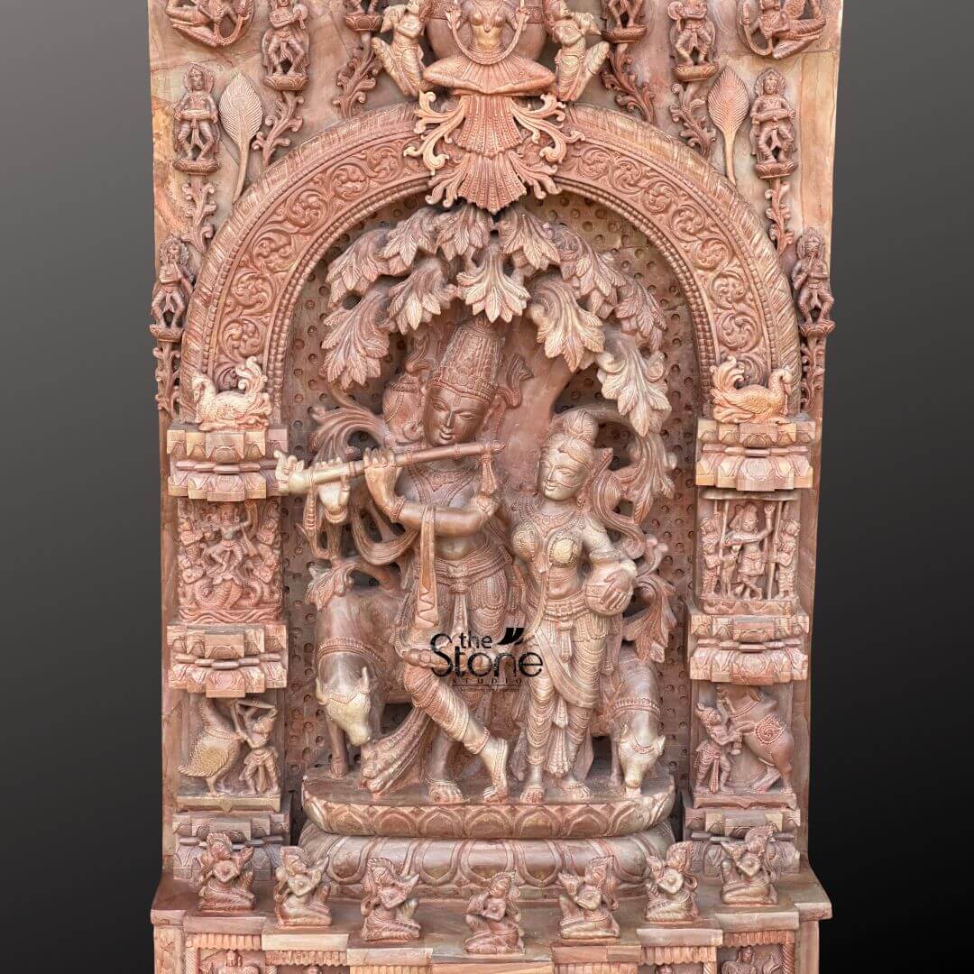 Pink Stone Radha Krishna Murti for Home 4ft 8 This close-up image showcases a beautiful Traditional Radha Krishna Idol in Pink Stone, carved as a 4-foot tall, highly detailed panel. The central and most prominent feature is the depiction of Lord Krishna playing his flute, standing gracefully with one leg crossed under a lush, intricately sculpted tree. Beside him, Radha stands in an attentive and loving posture, her figure also delicately carved with traditional attire and jewelry, all from the warm, natural pink stone. The carving is incredibly rich in detail, highlighting the classical artistry. Krishna's crown, the peacock feather adorning it, their expressions, and the folds of their garments are all rendered with precision. To Krishna's left, a cow is partially visible, adding to the idyllic pastoral setting often associated with Krishna. The main Radha Krishna scene is framed within a grand, deeply carved archway, which itself is adorned with intricate traditional motifs and smaller figures. The vertical side panels flanking this central arch are densely packed with numerous small sculptures, depicting devotees, celestial beings, and various mythological elements, creating a rich tapestry of narrative around the divine couple. The base below the main figures also features multiple tiers of finely carved friezes and smaller alcoves, each holding additional sculptures of figures and animals. This Traditional Radha Krishna Idol effectively captures the essence of devotion and classical Hindu artistry in a luxurious pink stone medium.