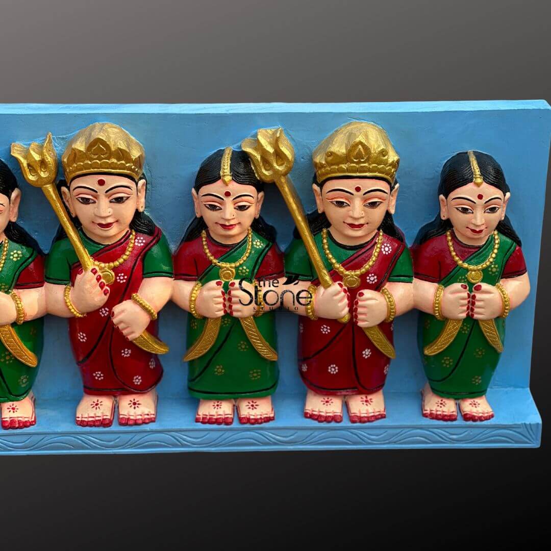 Sati Aasra Devi Statue 4ft 6 This close-up image displays a segment of a Traditional Sati Aasra Devi Sculpture, showcasing five of the identically styled female deities. The entire panel is 4 feet in length and features figures likely carved from black granite stone and then vibrantly painted. Each Devi figure is depicted with traditional, stylized features: round faces, large, dark eyes, and prominent red bindis on their foreheads. They are adorned with golden crowns and matching golden necklaces, with small earrings. Their traditional Indian garments alternate between rich red and deep green, each embellished with subtle, lighter-colored floral or star patterns. Their small, visible feet at the base are decorated with white floral motifs. The figures hold various divine attributes or gestures. Two of the visible figures are holding a golden trident (trishul) in one hand, symbolizing divine power and protection. The other three visible figures have their hands clasped together, suggesting a gesture of prayer or reverence. The entire ensemble is mounted on a solid, light blue rectangular panel, which acts as a contrasting backdrop and a sturdy base for the individually sculpted figures. The bold colors on the black granite stone base highlight the intricate details and traditional artistry, making this Sati Aasra Devi Sculpture a significant piece of devotional art.