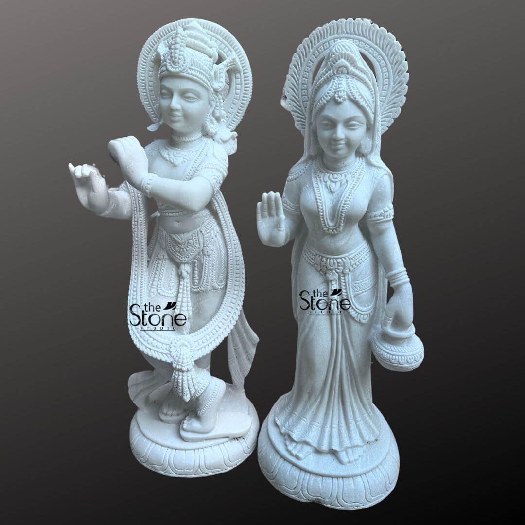 Radha Krishna Marble Stone Statue 3ft 4 This image presents a stunning Unique Radha Krishna Marble Idol featuring a pair of 3-foot tall white marble statues of Radha and Krishna. Expertly carved the deities are displayed against a dark neutral background allowing the pristine white marble and intricate details to stand out. On the left Lord Krishna stands in his iconic Tribhanga posture playing his flute. He is richly adorned with finely sculpted jewelry, a detailed crown with a peacock feather and flowing garments all rendered with exceptional precision in the white marble. A long garland also encircles his form. A prominent intricately carved circular halo is behind his head signifying his divine aura. On the right Radha stands gracefully her right hand raised in a gesture of blessing and her left hand holding a small pot or offering. She is also beautifully adorned with a crown, necklaces, bracelets, and a flowing sari, all intricately carved from the same white marble. A similar finely carved circular halo is behind her head. Both statues stand on individual ornate lotus pedestals which are integrated into the overall marble carving and feature additional carved details. The smooth, polished surface of the white marble statue reflects light softly emphasizing the delicate features, the extensive carvings on their attire and jewelry, and the divine expressions of Radha and Krishna. This Unique Radha Krishna Marble Idol embodies sacred love and traditional Indian artistry in a captivating and distinct manner.