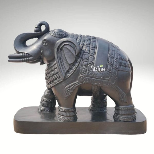 An exquisitely hand-carved 21-inch blackstone elephant statue, masterfully designed to serve as a powerful Vastu symbol for any home or office. The sculpture depicts a majestic elephant in a stable standing pose with its trunk curved upwards and back, a traditional gesture representing victory, good luck, and prosperity according to Vastu Shastra. The elephant is adorned with an ornate decorative blanket across its back, featuring intricate geometric and floral carvings. Matching ornamental bands wrap around its legs and neck, showcasing the depth of traditional Indian stone craftsmanship. The deep charcoal hue and polished finish of the natural blackstone give the statue a sophisticated, rustic appeal. Standing on a solid, integrated rectangular pedestal, this Vastu elephant statue in black acts as a guardian figure, perfect for placement in a home foyer, living room, or as a centerpiece in a garden landscape to invite groundedness, protection, and auspicious energy.