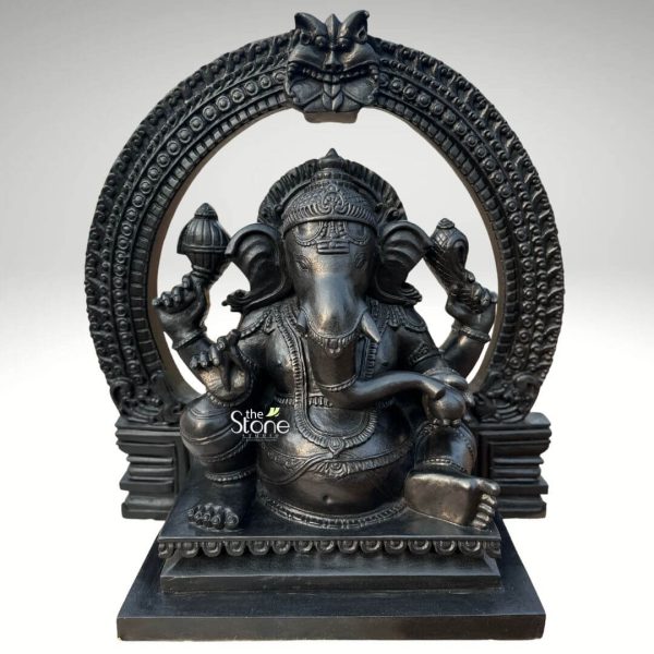 An intricately detailed 24-inch Vighnaharta Ganesh idol, masterfully crafted from high-quality fibre with a sophisticated black stone finish. This 2-foot spiritual sculpture captures the deity in a stable and majestic seated pose, making it an ideal centerpiece for a home mandir, office, or meditation space. The idol features a rich, matte charcoal black texture that mimics the appearance of traditional blackstone carvings. Lord Ganesha is depicted with four arms, holding sacred spiritual symbols such as the discus and conch shell, while his large expressive ears and gracefully curved trunk symbolize wisdom and the ability to listen. He is beautifully adorned with a towering tiered crown (mukut), layered necklaces, and traditional armlets. A grand, circular Prabhavali (ornate arch) frames the deity, featuring deep textures and a protective Kirtimukha face at the peak, which adds a sense of ancient temple grandeur. The entire Ganesha fibre statue rests on a solid rectangular pedestal embellished with subtle lotus petal relief carvings. This durable and lightweight sculpture serves as a powerful symbol of the remover of obstacles, inviting peace, prosperity, and divine protection into any environment.
