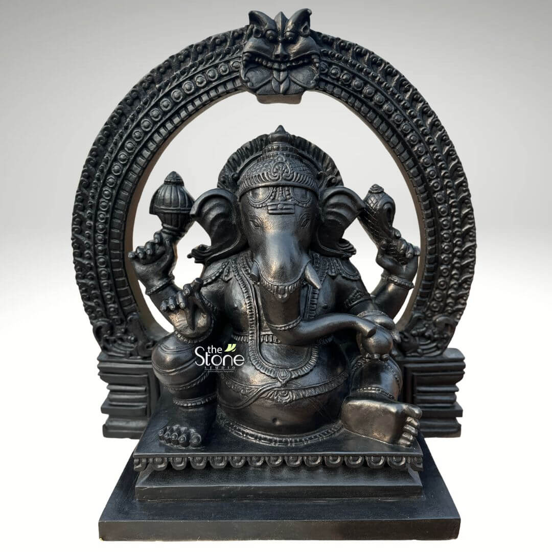 Vighnaharta Ganesh Idol 2ft 1 An intricately detailed 24-inch Vighnaharta Ganesh idol, masterfully crafted from high-quality fibre with a sophisticated black stone finish. This 2-foot spiritual sculpture captures the deity in a stable and majestic seated pose, making it an ideal centerpiece for a home mandir, office, or meditation space. The idol features a rich, matte charcoal black texture that mimics the appearance of traditional blackstone carvings. Lord Ganesha is depicted with four arms, holding sacred spiritual symbols such as the discus and conch shell, while his large expressive ears and gracefully curved trunk symbolize wisdom and the ability to listen. He is beautifully adorned with a towering tiered crown (mukut), layered necklaces, and traditional armlets. A grand, circular Prabhavali (ornate arch) frames the deity, featuring deep textures and a protective Kirtimukha face at the peak, which adds a sense of ancient temple grandeur. The entire Ganesha fibre statue rests on a solid rectangular pedestal embellished with subtle lotus petal relief carvings. This durable and lightweight sculpture serves as a powerful symbol of the remover of obstacles, inviting peace, prosperity, and divine protection into any environment.