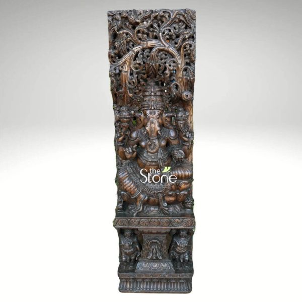 This image showcases a magnificent 5-foot tall Wood Ganesha Wall Panel intricately carved from dark wood with an extraordinary level of detail. The panel presents Lord Ganesha as the central, seated figure surrounded by a rich tapestry of carved motifs and a sprawling tree design. Lord Ganesha is depicted in a traditional relaxed sitting posture with his multiple arms holding his characteristic attributes, including a modak (sweet) in one of his lower hands. He is richly adorned with finely sculpted jewelry, a crown, and an ornate dhoti, all rendered with deep, precise cuts that give the wood carving remarkable texture and three-dimensionality. His trunk curves gracefully to his left. Above Ganesha, an elaborate tree motif forms a magnificent canopy, with countless branches and leaves carved in high relief, creating a sense of natural grandeur. The entire background of the panel, especially the upper section and sides, is densely filled with intricate floral patterns, scrollwork, and possibly smaller divine or animal figures, showcasing the meticulous craftsmanship. The wooden Ganesha panel stands on a robust, multi-tiered wooden pedestal, which is also intricately carved with traditional designs, including figures that appear to be guardian lions or mythical creatures at its base. The dark wood has a rich, deep tone, and the texture of the carving is highlighted by the lighting, emphasizing the artisanal skill involved. This wood Ganesha wall panel is a grand and spiritually significant piece, perfect for a prominent display as a work of art and devotion.