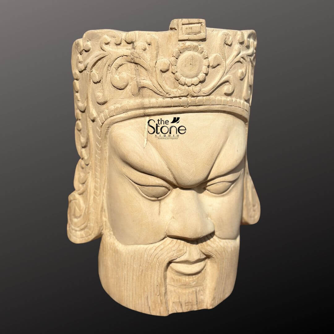 Royal Head Sculpture 12" 3 This image features a detailed 12-inch Wooden Head Sculpture carved from light-colored wood, presented against a dark, neutral background. The sculpture depicts a male face with a stern and commanding expression, possibly representing a warrior, king, or deity from East Asian or ancient cultures. The face has strong, defined features: deeply furrowed brows, intense, narrow eyes that gaze downward, a prominent straight nose, and a closed mouth with a full, sculpted beard and mustache. The expression conveys a sense of power, authority, and perhaps a subtle hint of fierce determination. Crowning the head is an elaborate headdress or helmet, richly detailed with intricate floral patterns, swirling motifs, and a central circular emblem, possibly a sun or a stylized flower. The precision of the carving is evident in every element, with all features meticulously shaped from the single piece of wood. The natural grain of the wood is visible throughout the sculpture, particularly on the face and beard, adding to its authentic texture and handcrafted appeal. This wooden head sculpture serves as an impactful and artistic focal point, blending historical aesthetics with natural material.