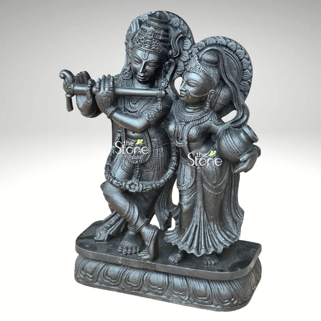 2ft Radha Krishna Black Stone Statue 2 A striking 2-feet black stone Radha Krishna idol is captured in a slightly elevated frontal shot against a plain light background. The statue depicts Lord Krishna and Radha standing side-by-side on a decorative lotus-patterned base. Lord Krishna is positioned on the left, playing a flute held to his lips with one leg crossed over the other in his characteristic pose. He is adorned with an elaborate crown intricate jewelry and flowing garments all carved with fine detail. Radha stands gracefully to his right looking lovingly towards Krishna. She carries a small pot or kalash in her left hand and is also richly decorated with a crown, jewelry, and draped attire. Both figures are carved from dark black stone, which gives the statue a profound and traditional appearance with the details highlighted by subtle variations in light. A decorative halo or arch behind Krishna's head further emphasizes their divine presence. This detailed image beautifully conveys the devotion and artistry of the black stone Radha Krishna statue, making it highly relevant to an article about such idols.