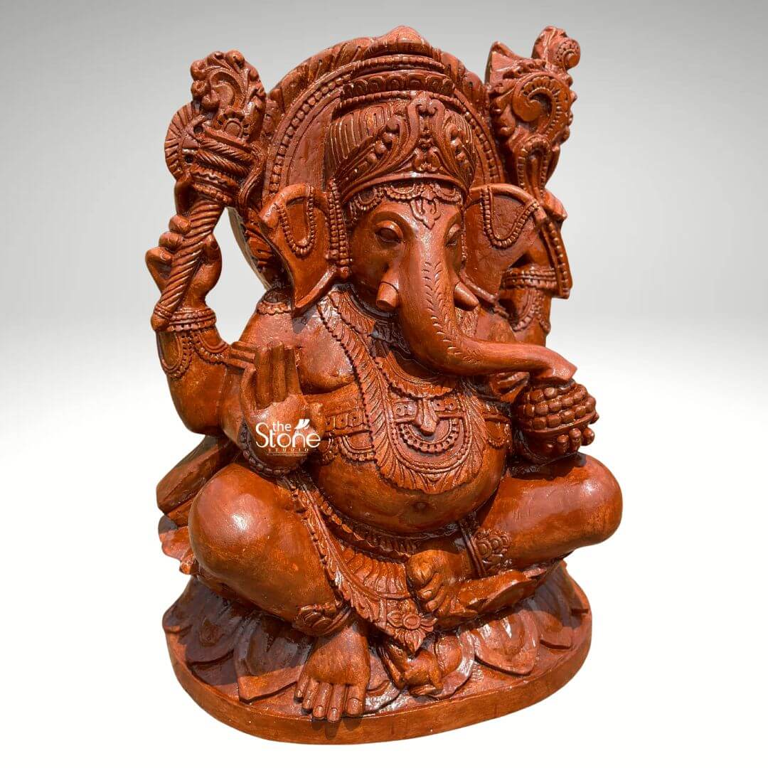 Pillaiyar Ganesha Statue 2.5ft 2 A magnificent 2.5-feet sandstone Vinayagar statue is presented from a frontal, slightly elevated perspective against a plain light background. The statue is carved from reddish brown sandstone showcasing its natural texture and warm earthy tones. Lord Ganesha is depicted in a traditional serene seated posture cross-legged on an intricately carved lotus base. He has four arms his upper right hand holds an axe or goad while his upper left hand grasps a noose. His lower right hand is raised in a gesture of blessing and his lower left hand holds a bowl of modaks with his trunk curving towards it. Ganesha is richly adorned with an elaborate crown intricate necklaces armlets, and a sacred thread draped across his plump belly, which symbolizes prosperity. His large, fan-like ears and expressive eyes are finely detailed. A magnificent halo or prabhavali, with a detailed outer edge and internal carvings, frames his head, emphasizing his divine stature. The textured surface of the sandstone highlights the intricate details of all the carvings, from the delicate features of Ganesha to the elaborate motifs on the backdrop and base. This detailed image powerfully conveys the spiritual majesty and artistic craftsmanship of the sandstone Vinayagar statue, making it highly relevant to an article discussing stone idols.