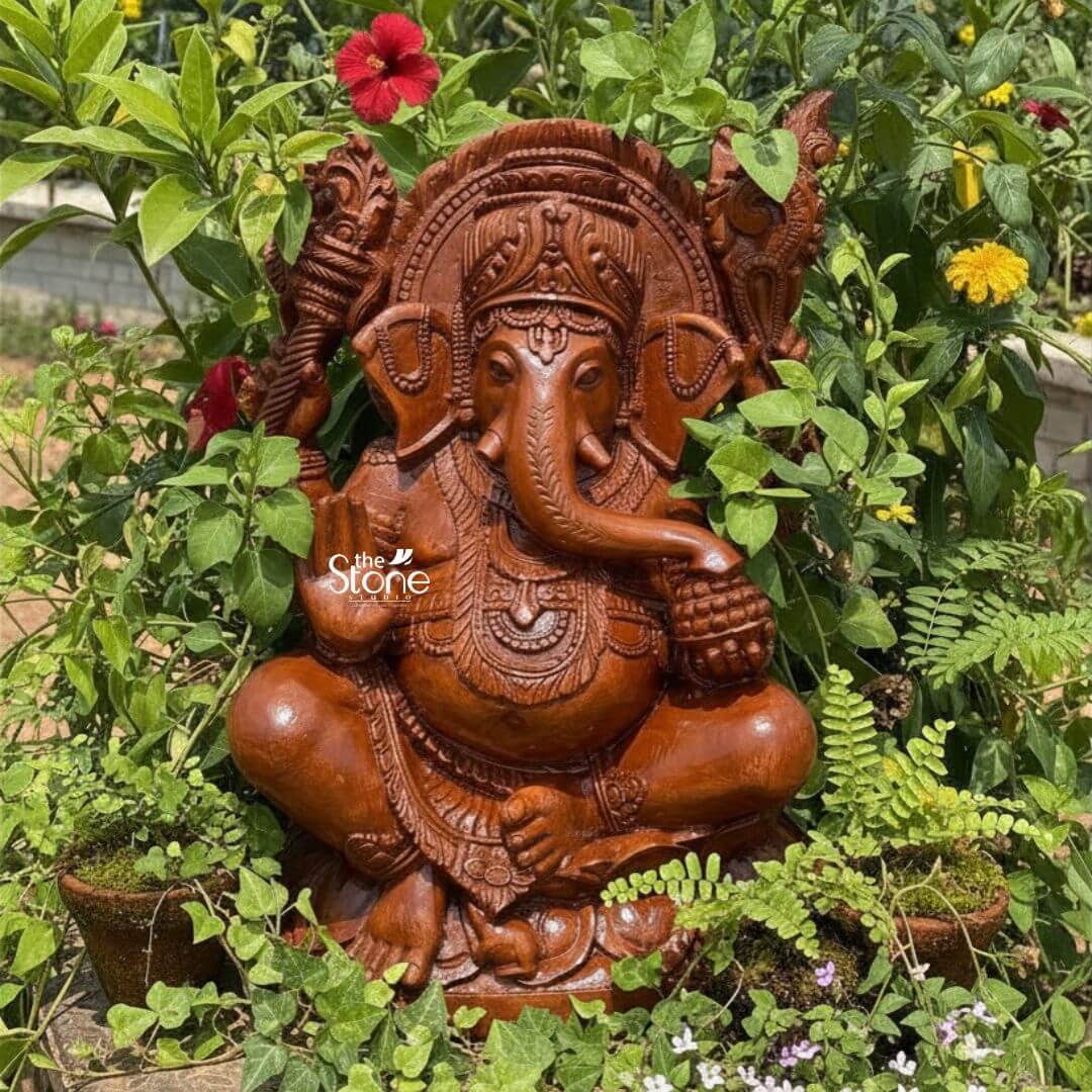 Pillaiyar Ganesha Statue 2.5ft 3 A magnificent 2.5-feet sandstone Vinayaka statue is beautifully nestled in a lush outdoor garden setting surrounded by vibrant green plants and colorful flowers. The statue is carved from reddish-brown sandstone showcasing its rich natural texture and warm earthy tones, which blend harmoniously with the botanical environment. Lord Ganesha is depicted in a traditional, serene seated posture, cross-legged, on an intricately carved lotus base, radiating an aura of wisdom, prosperity, and benevolence. Ganesha has four arms; his upper right hand holds an axe or goad, while his upper left hand grasps a noose. His lower right hand is raised in a gesture of blessing and his lower left hand holds a bowl of modaks with his trunk curving towards it. He is richly adorned with an elaborate crown, intricate necklaces, armlets, and a sacred thread draped across his plump belly. His large fan-like ears and expressive eyes are finely detailed. A magnificent halo or prabhavali, with a detailed outer edge and internal carvings, frames his head, emphasizing his divine stature. The textured surface of the sandstone highlights the intricate details of all the carvings. The surrounding greenery includes various leafy plants, small potted ferns, and pops of color from red and yellow flowers, creating a serene and natural backdrop. This detailed image powerfully conveys the spiritual majesty and artistic craftsmanship of the sandstone Vinayaka statue, making it highly relevant to an article discussing such revered idols.