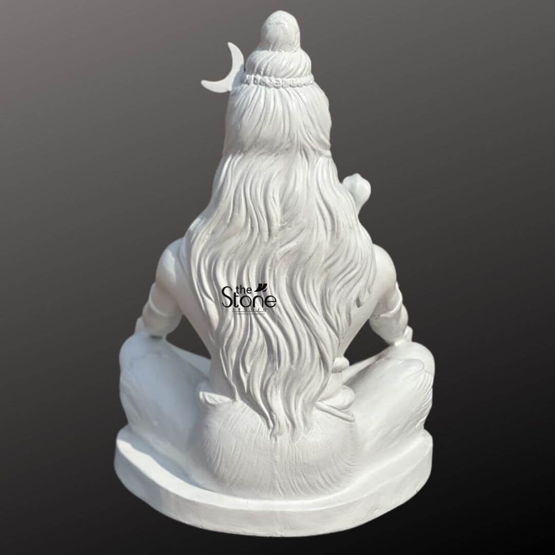 2ft Shiva Meditation Statue 7 A serene 2-foot fiber Lord Shiva statue ideal for home worship is presented from a slightly elevated frontal perspective against a plain dark background. The statue is entirely white embodying purity and calm. Lord Shiva is depicted in a meditative seated posture (Padmasana or Siddhasana), with his legs crossed and hands resting open in his lap, likely in Dhyana Mudra. His eyes are gently closed or downcast, conveying deep introspection and tranquility. Shiva is adorned with his traditional attributes: a crescent moon is nestled in his matted hair (Jata), from which the Ganges river is often said to flow (represented by flowing locks). A cobra is draped around his neck and shoulders, and he wears rudraksha bead necklaces and armlets. His third eye is subtly marked on his forehead. The smooth finish of the fiber material highlights the anatomical details and the delicate sculpting of his features and adornments. The statue rests on a simple, oval-shaped white base. This detailed image effectively conveys the spiritual essence and artistic craftsmanship of the fiber Shiva statue, making it highly relevant to an article discussing home idols.