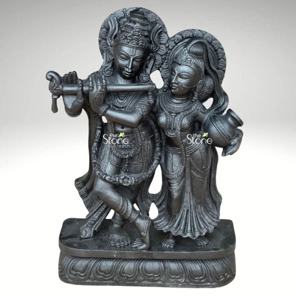 A captivating 2-feet black stone Radha Krishna statue is showcased in a slightly elevated frontal view against a plain light background. The sculpture beautifully portrays Lord Krishna and Radha standing together on an ornate lotus-patterned rectangular base. Lord Krishna is positioned on the left elegantly playing his flute which he holds to his lips. His posture is dynamic with one leg crossed and he is richly adorned with an elaborate crown intricate necklaces and finely carved garments. Radha stands gracefully to his right her gaze directed towards Krishna embodying devotion. She holds a traditional pot or kalash in her left hand and is equally adorned with a crown, jewelry, and flowing attire, all meticulously carved. Both divine figures are rendered in deep black stone giving the statue a powerful and classic aesthetic, with the sculptural details accentuated by the lighting. A subtle halo or decorative element rises behind Krishna's head, completing the divine representation. This detailed image vividly conveys the spiritual essence and artistic craftsmanship of the 2-foot black stone Radha Krishna statue, making it highly relevant to an article focused on stone idols.