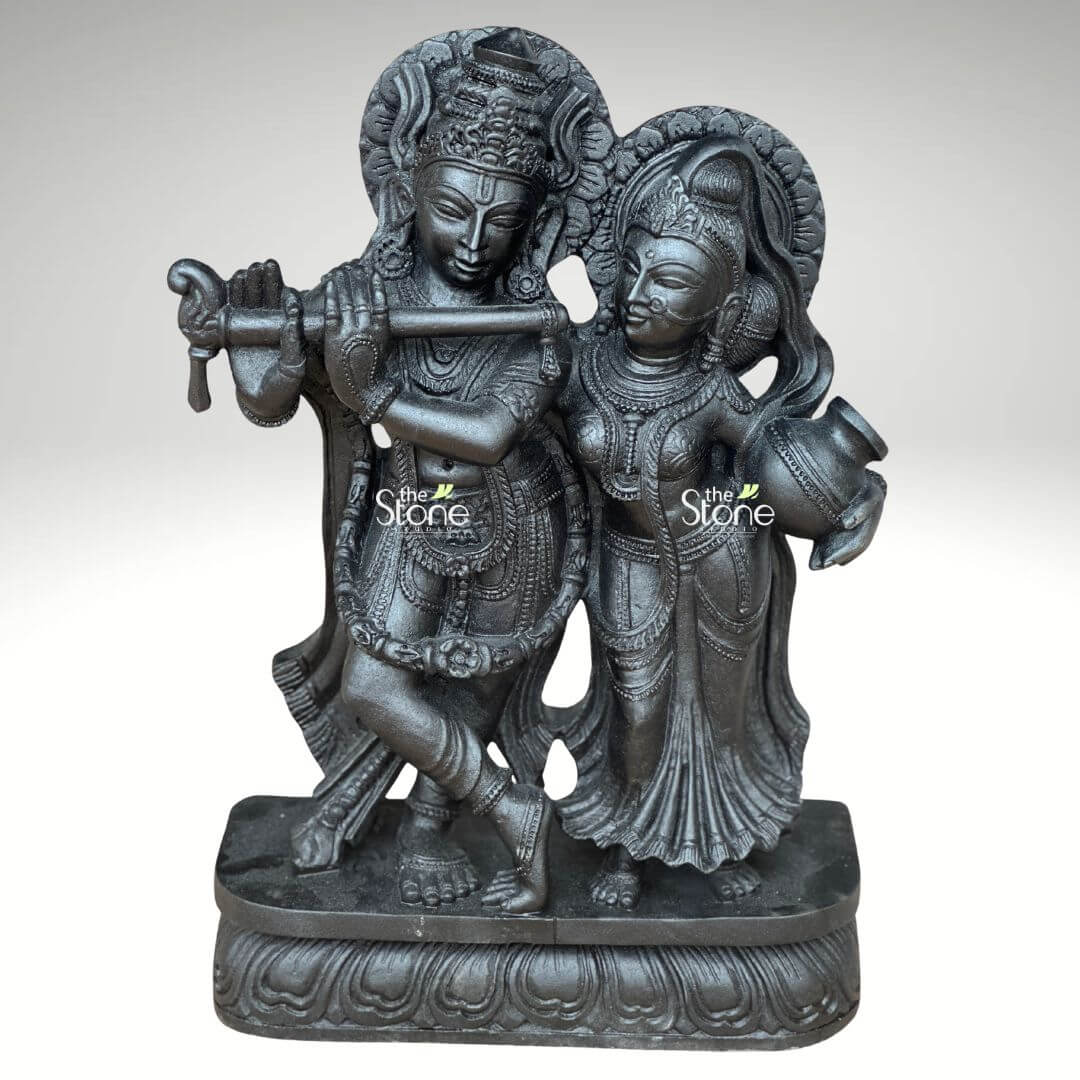 2ft Radha Krishna Black Stone Statue 1 A captivating 2-feet black stone Radha Krishna statue is showcased in a slightly elevated frontal view against a plain light background. The sculpture beautifully portrays Lord Krishna and Radha standing together on an ornate lotus-patterned rectangular base. Lord Krishna is positioned on the left elegantly playing his flute which he holds to his lips. His posture is dynamic with one leg crossed and he is richly adorned with an elaborate crown intricate necklaces and finely carved garments. Radha stands gracefully to his right her gaze directed towards Krishna embodying devotion. She holds a traditional pot or kalash in her left hand and is equally adorned with a crown, jewelry, and flowing attire, all meticulously carved. Both divine figures are rendered in deep black stone giving the statue a powerful and classic aesthetic, with the sculptural details accentuated by the lighting. A subtle halo or decorative element rises behind Krishna's head, completing the divine representation. This detailed image vividly conveys the spiritual essence and artistic craftsmanship of the 2-foot black stone Radha Krishna statue, making it highly relevant to an article focused on stone idols.