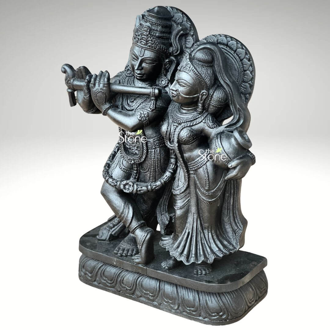 2ft Radha Krishna Black Stone Statue 5 A captivating 2-feet black stone Radha Krishna statue is showcased in a slightly elevated frontal view against a plain light background. The sculpture beautifully portrays Lord Krishna and Radha standing together on an ornate lotus-patterned rectangular base. Lord Krishna is positioned on the left elegantly playing his flute which he holds to his lips. His posture is dynamic, with one leg crossed and he is richly adorned with an elaborate crown intricate necklaces and finely carved garments. Radha stands gracefully to his right, her gaze directed towards Krishna embodying devotion. She holds a traditional pot or kalash in her left hand and is equally adorned with a crown, jewelry, and flowing attire, all meticulously carved. Both divine figures are rendered in deep black stone giving the statue a powerful and classic aesthetic, with the sculptural details accentuated by the lighting. A subtle halo or decorative element rises behind Krishna's head completing the divine representation. This detailed image vividly conveys the spiritual essence and artistic craftsmanship of the 2-foot black stone Radha Krishna statue making it highly relevant to an article focused on stone idols.