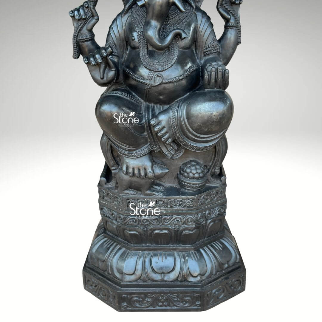 3.5ft Ganesha Garden Statue 4 A close-up eye-level shot captures the lower body and intricate details of a magnificent 3.5-foot black stone Ganesha statue perfectly suited as an outdoor centerpiece. The focus is on Ganesha's substantial form seated in a meditative posture with his legs crossed resting upon an elaborately carved lotus pedestal. His left hand is prominently featured holding a bowl of modak, symbolizing abundance and wisdom. The statue's robust belly is visible adorned with delicate jewelry that emphasizes the smooth dark texture of the black stone. The pedestal itself is a work of art featuring detailed floral and geometric patterns and resting on a sturdy multi-tiered octagonal base with elegant lotus petal carvings. The fine craftsmanship extends to Ganesha's feet and the textures of his drapery all rendered with precision. This perspective highlights the statue's impressive scale and the exquisite detail of its lower half reinforcing its suitability as a 3.5ft Ganesha Outdoor Statue designed for durability and aesthetic appeal in any open-air environment.