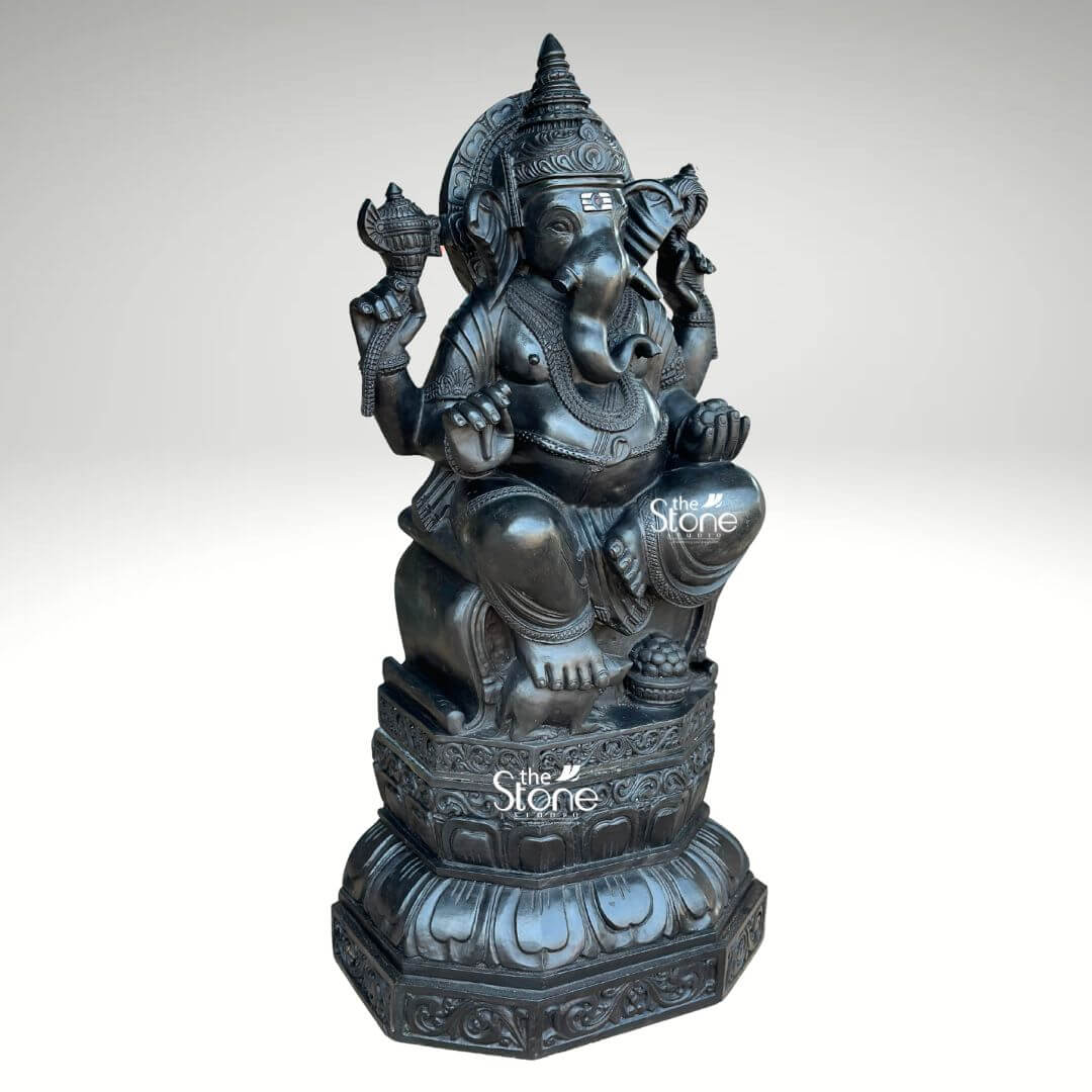 3.5ft Ganesha Garden Statue 2 This compelling image displays a grand 3.5-foot Lord Ganesha stone statue crafted from exquisite black stone positioned centrally against a muted background that emphasizes its intricate details and commanding presence. Ganesha is depicted in a traditional seated posture radiating a sense of profound tranquility and wisdom. His iconic elephant head is meticulously sculpted featuring large gracefully fanned ears, deep-set intelligent eyes and a trunk elegantly curled to his left reaching towards a symbolic offering. The deity is richly adorned with an elaborate tiered crown multiple necklaces and detailed armlets and anklets all carved with remarkable precision that captures the textural nuances of the black stone. The statue is anchored to a beautifully designed multi-layered lotus pedestal, a revered symbol of purity and spiritual awakening in Hindu iconography. This pedestal, in turn rests upon a robust octagonal base further embellished with ornate carvings of lotus petals and intricate floral patterns reinforcing the sacred and artistic value of the piece. In his upper right hand Ganesha holds a conch shell and in his upper left a finely carved axe – traditional symbols that represent divine sound, the breaking of obstacles and the discernment of truth. His lower right hand is raised in a gesture of Abhaya Mudra offering blessings and assurance while his lower left hand cradles a bowl of modak, his favorite sweet, symbolizing the sweet rewards of spiritual devotion and prosperity. The smooth polished surface of the black stone enhances the statue's majestic quality, giving it a timeless and sophisticated appeal. Every curve and contour of the 3.5ft Lord Ganesha Stone Statue is rendered with expert craftsmanship making it not just a religious icon but also a significant work of art. This detailed alt text ensures that visually impaired users receive a comprehensive understanding of the statue's appearance, its cultural significance and its connection to the article's focus on a substantial and beautifully crafted Ganesha stone sculpture.