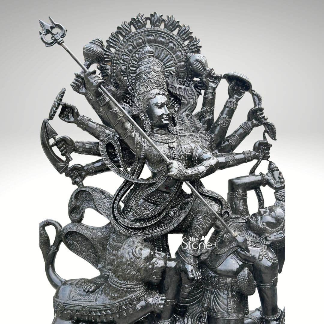6ft Mahishasura Mardini Durga Statue 2 An exquisitely detailed close-up of a 6-foot tall Durga Mahishasura Mardini black stone idol masterfully hand-carved from premium natural black stone. This large-scale spiritual masterpiece captures the Hindu Goddess in her most powerful form representing the triumph of good over evil as she slays the buffalo demon, Mahishasura. Goddess Durga is depicted as Dashabhuja with her hands radiating in a fan-like pattern, each holding a sacred weapon of divine power. Her central hands are shown thrusting a long, slender trident trishula into the demon. The deity features a serene yet determined facial expression, adorned with a towering, tiered mukut and layered temple jewelry that showcases exceptional artisan craftsmanship. The sculpture is framed by a grand, circular Prabhavali featuring deeply carved floral and flame-like relief patterns. At the base, her fierce lion vehicle vahana is depicted attacking the demon, who is shown in human form emerging from the buffalo. The refined polished finish of the black stone Durga statue emphasizes the intricate textures of the carvings and the realistic folds of her traditional attire. This monumental 6ft Durga murti serves as a majestic spiritual centerpiece, ideal for a grand temple sanctuary, luxury foyer, or a dedicated prayer space, designed to invite protection and divine energy.