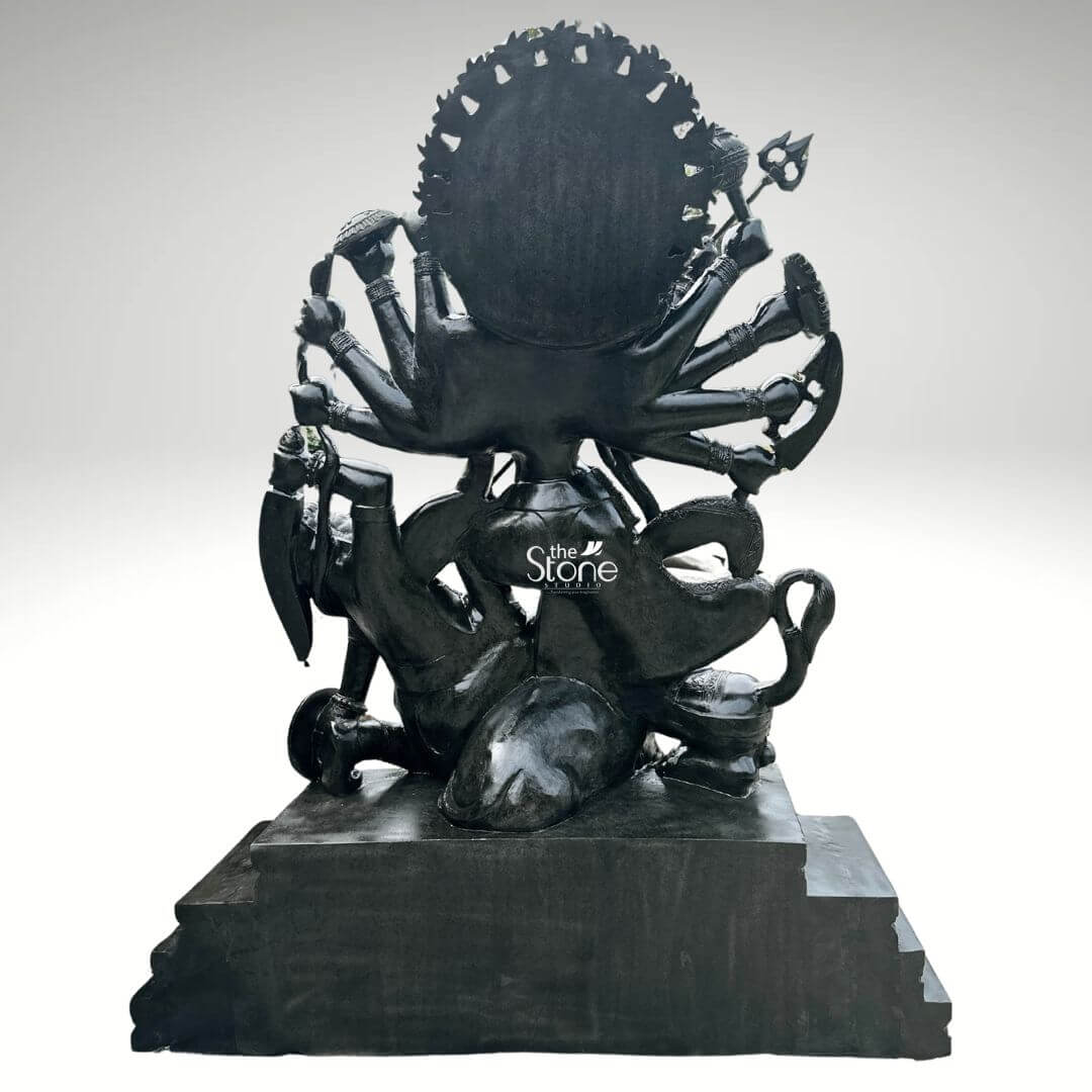 6ft Mahishasura Mardini Durga Statue 6 A rear view of a majestic 6-foot tall Durga Mahishasura Mardini idol masterfully hand-carved from premium natural black stone. This perspective highlights the exceptional 360-degree craftsmanship of the monumental piece, intended as a grand spiritual centerpiece for a temple sanctuary. The sculpture depicts the ten-armed Goddess Durga in a dynamic battle stance, standing over the buffalo demon, Mahishasura. From the back, her ten arms are seen radiating outward in a fan-like pattern, each holding sacred weapons representing divine strength. The intricate detailing extends to the deity’s flowing hair and the realistic fabric folds of her traditional attire. A grand circular Prabhavali with flame-edged relief carvings frames her head, symbolizing divine power. The entire black stone Durga statue is anchored by a sturdy, multi-tiered rectangular base. The refined, polished charcoal finish emphasizes the heavy and permanent nature of the stone, illustrating its durability for a temple foyer or a large outdoor spiritual landscape. This large-scale Mahishasura Mardini sculpture perfectly captures the essence of traditional Indian stone masonry, designed to invite an aura of protection and divine triumph into any sacred environment.