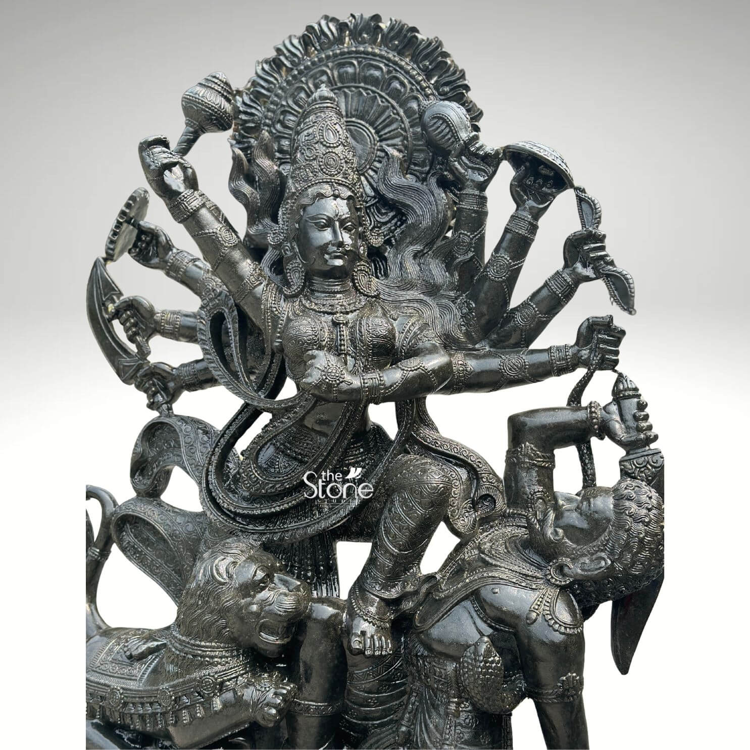 6ft Mahishasura Mardini Durga Statue 4 A majestic 6-foot tall Durga Mahishasura Mardini statue masterfully hand-carved from premium natural black stone with a refined polished charcoal finish. This close-up highlights the exceptional Indian stone masonry applied to the Goddess in her ten-armed form. She is captured in the iconic and powerful moment of divine triumph slaying the buffalo demon Mahishasura to restore cosmic balance. The deity is depicted with a serene yet determined facial expression holding various divine weapons including a long trishula sword, and discus—in her radiating arms. She is adorned with an elaborate, towering mukut layered temple necklaces, and ornate armlets. Goddess Durga stands in a dynamic posture with one foot resting on her fierce lion vahana and the other firmly placed on the demon. Framing her head is a grand, circular Prabhavali featuring intricate rhythmic relief patterns. Every detail, from the realistic, fluid fabric folds of her traditional attire to the textured mane of the lion, emphasizes the masterful craftsmanship of this large-scale piece. This 6ft blackstone Durga idol serves as a profound spiritual centerpiece, ideal for a grand temple sanctuary, a luxury residential foyer, or a dedicated meditation space designed to invite protection and divine energy.