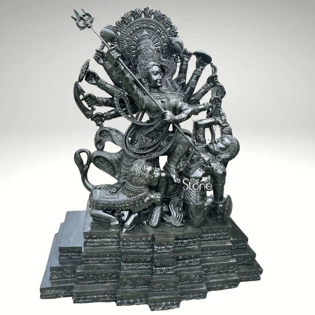 6ft Mahishasura Mardini Durga Statue 5 A majestic 6-foot tall Lord Durga statue, masterfully hand-carved from premium natural black stone serving as a grand spiritual centerpiece for a temple foyer or a luxury landscape. This monumental 6ft blackstone Durga idol captures the powerful form of Mahishasura Mardini the slayer of the buffalo demon symbolizing the ultimate triumph of good over evil. The Goddess is depicted in a dynamic warrior posture with ten arms radiating in a fan-like pattern, each hand holding a sacred weapon of divine power. Her central hands are shown thrusting a long, slender trident trishula into the demon Mahishasura, who is depicted in human form emerging from the buffalo. Goddess Durga features a serene yet determined facial expression and is adorned with a towering, tiered mukut and layered temple jewelry that showcases exceptional artisan craftsmanship. The sculpture is framed by a grand, circular Prabhavali featuring deeply carved traditional floral and flame-like relief patterns. At her feet, her fierce lion vehicle participates in the battle, with every detail of its mane and muscles rendered in fine detail. The entire 6-foot black stone sculpture rests on a massive multi-tiered stepped pedestal embellished with rhythmic architectural patterns. The refined polished charcoal finish of the stone highlights the rich heritage of traditional Indian stone masonry making this large-scale Durga Murti a powerful symbol of strength, protection, and divine energy for any prominent residential or religious space.