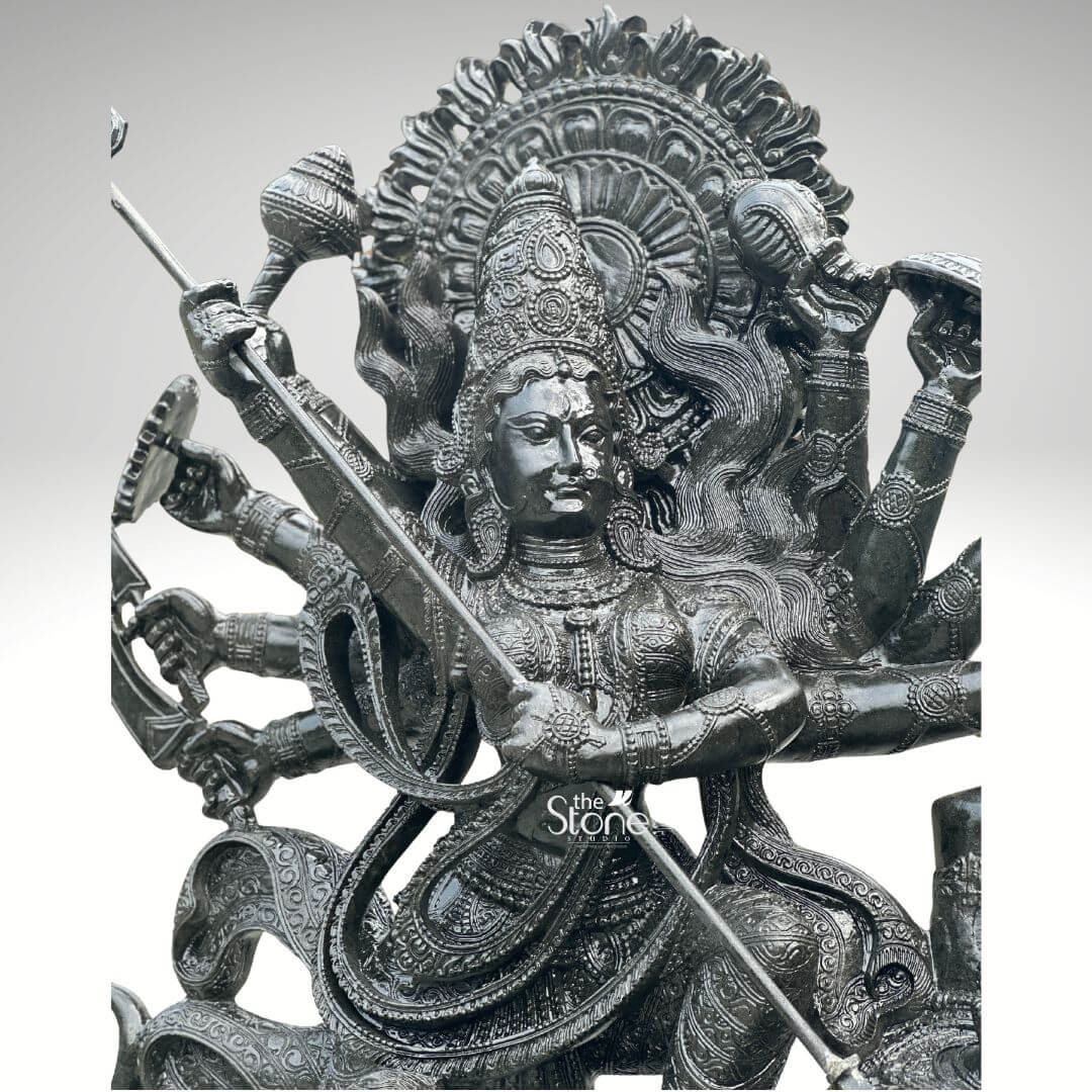 6ft Mahishasura Mardini Durga Statue 8 An exquisite close-up of a 6-foot tall Goddess Durga Mahishasura Mardini statue masterfully hand-carved from premium natural black stone. This detailed shot highlights the superior craftsmanship of the blackstone Durga idol capturing her in the powerful role of the slayer of the buffalo demon. The Goddess features a serene yet determined facial expression, adorned with a traditional nose ring and an intricately carved tiered crown mukut. Framing her head is a grand circular Prabhavali embellished with rhythmic, sunburst-like relief patterns that symbolize her divine aura. As the ten-armed Dashabhuja Durga several of her hands are shown radiating outward, each holding a sacred weapon, while her primary hands firmly grasp a long, slender trident trishula used to restore cosmic order. The sculpture showcases masterful Indian stone masonry visible in the finely detailed temple jewelry, layered necklaces, and the realistic, fluid folds of her traditional attire etched into the dark, polished stone. This monumental 6ft Durga murti serves as a majestic spiritual centerpiece, ideal for a grand temple foyer, luxury villa entrance, or meditation sanctuary, designed to invite strength, protection, and divine energy into the space.