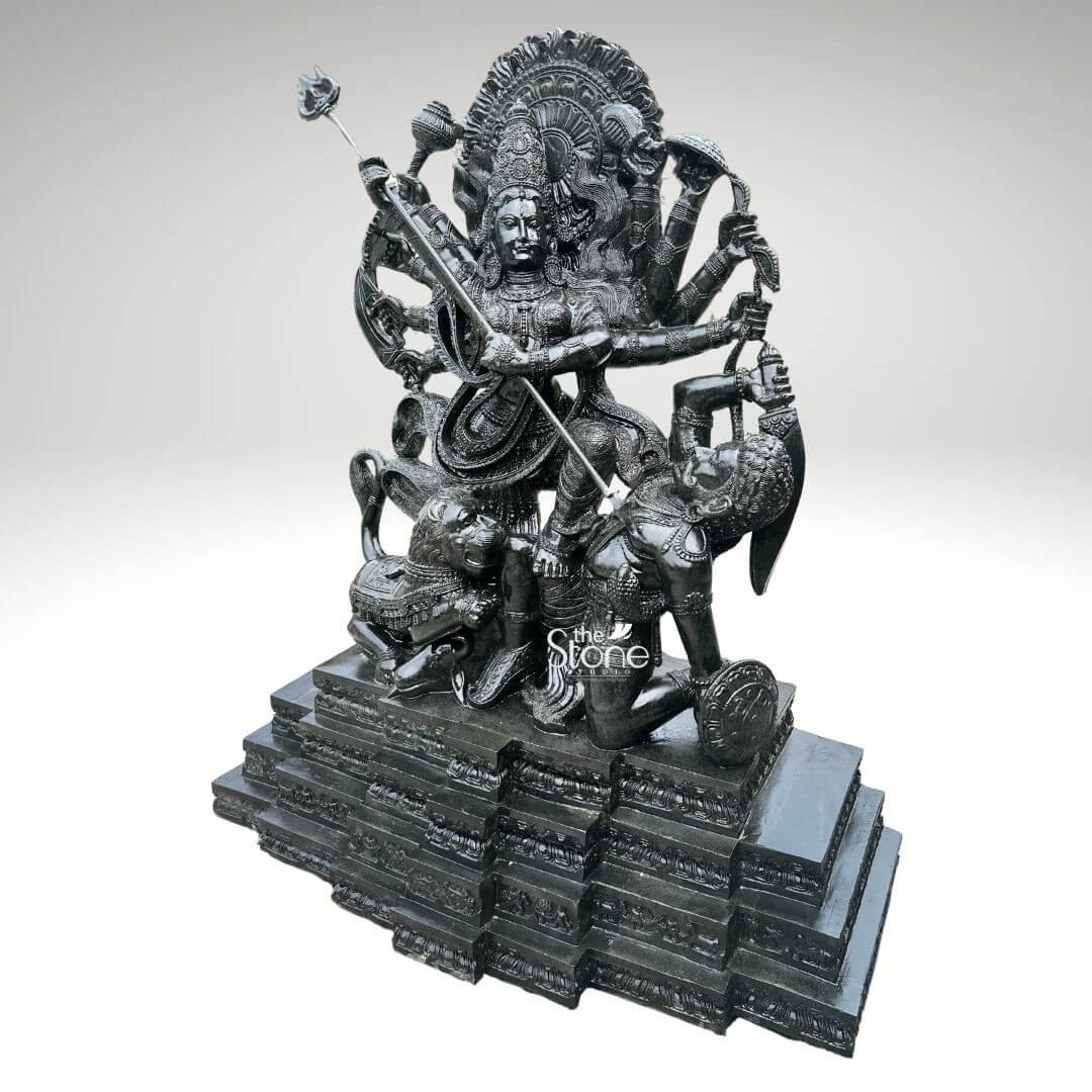 6ft Mahishasura Mardini Durga Statue 9 A monumental 6-foot tall Mahishasura Mardini Durga idol masterfully hand-carved from premium natural black stone serves as a majestic spiritual centerpiece for a temple foyer or a luxury outdoor landscape. This large-scale black stone Durga statue captures the Goddess in her powerful ten-armed form, depicting the iconic moment of divine triumph as she slays the buffalo demon, Mahishasura. Seen from a three-quarter perspective to highlight its monumental scale and intricate depth, the deity is shown in a dynamic battle pose. Her central hands are shown thrusting a long, slender trident (trishula) into the demon, who is depicted in human form emerging from the buffalo. Goddess Durga features a serene yet determined facial expression and is adorned with a towering, tiered mukut and layered temple jewelry that showcases exceptional Indian stone masonry. The sculpture is framed by a grand, circular Prabhavali featuring deeply carved traditional floral and flame-like relief patterns. At her feet, her fierce lion vehicle participates in the battle with every detail of its mane and posture rendered with precision. The entire 6-foot black stone sculpture rests on a massive, multi-tiered stepped pedestal. The refined polished charcoal finish of the stone highlights the rich heritage of traditional craftsmanship, making this large-scale Durga Murti a powerful symbol of strength, protection, and divine energy for any prominent residential or religious space.