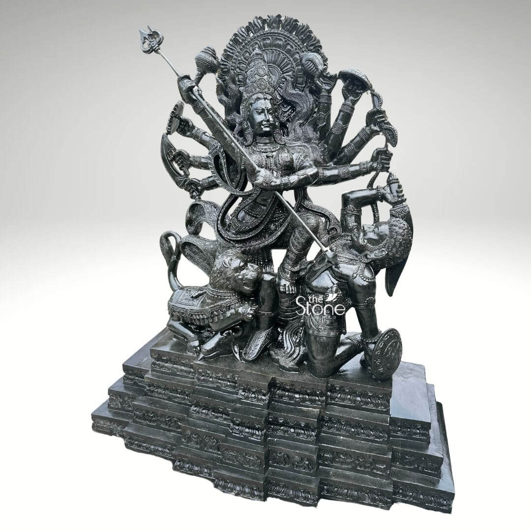 6ft Mahishasura Mardini Durga Statue 10 A magnificent 6-foot tall blackstone sculpture of Mahishasura Mardini Durga stands as the centerpiece. The multi-armed goddess Durga is depicted in a dynamic and powerful pose, with one foot on the ground and the other raised, poised to strike. She wields numerous weapons in her many hands, including a trident, bow and arrow, symbolizing her strength and divine power. Below Durga, the demon Mahishasura, transformed into a buffalo-demon, is subdued. The sculpture intricately captures the dramatic moment of their epic battle, showcasing Durga's triumph over evil. A lion, Durga's vahana is also carved beside her, adding to the grandeur of the composition. The entire sculpture rests on a multi-tiered, ornate pedestal, also crafted from blackstone, featuring intricate carvings that enhance its traditional aesthetic. The dark, polished stone gives the statue a commanding presence, highlighting the detailed craftsmanship and the fierce yet divine expression of the goddess. This sculpture is designed for a temple, embodying spiritual significance and artistic mastery.