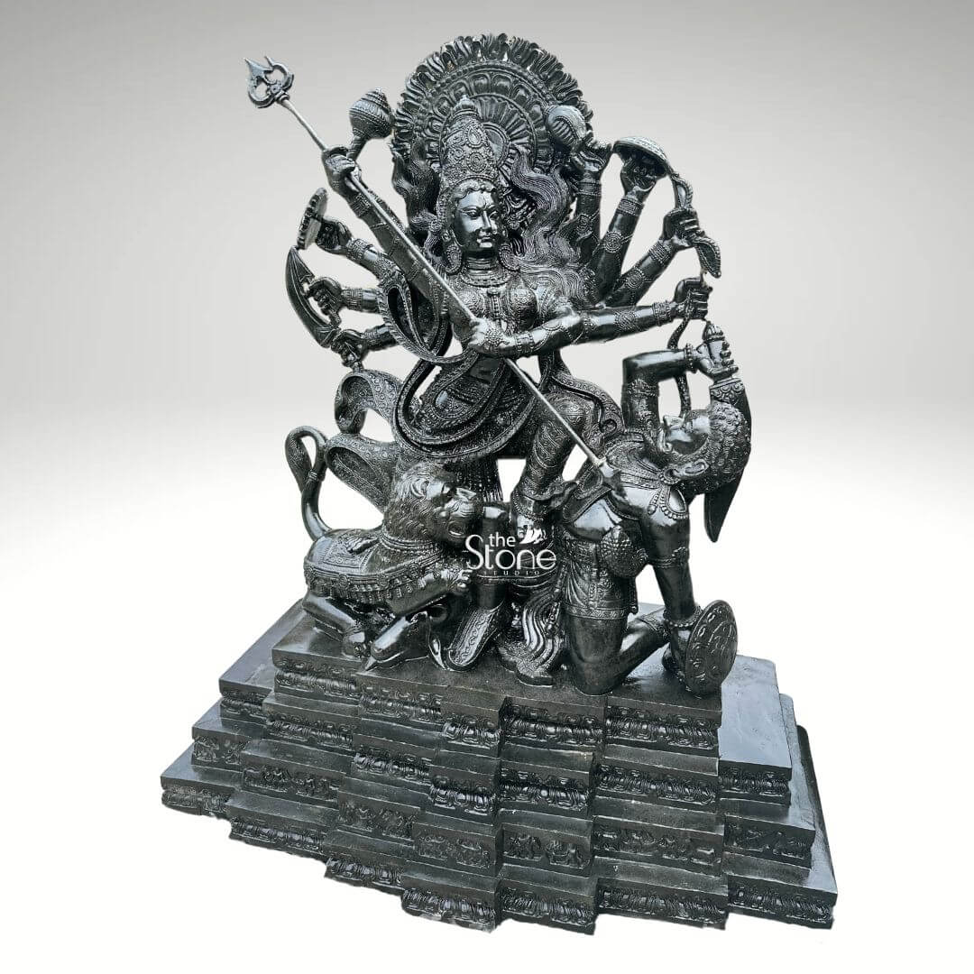 6ft Mahishasura Mardini Durga Statue 11 A monumental 6-foot tall Mahishasura Mardini idol masterfully hand-carved from premium natural black stone serves as a majestic spiritual centerpiece for a temple foyer or a luxury outdoor landscape. This large-scale black stone Durga statue captures the Goddess in her powerful ten-armed Dashabhuja form, depicting the iconic moment of divine triumph as she slays the buffalo demon, Mahishasura. Seen from a three-quarter perspective to highlight its monumental scale and intricate depth, the deity is shown in a dynamic warrior pose. Her central hands are shown thrusting a long, slender trident trishula into the demon, who is depicted in human form emerging from the buffalo. Goddess Durga features a serene yet determined facial expression and is adorned with a towering, tiered mukut and layered temple jewelry that showcases exceptional Indian stone masonry. The sculpture is framed by a grand, circular Prabhavali featuring deeply carved traditional floral and flame-like relief patterns. At her feet, her fierce lion vehicle participates in the battle, with every detail of its mane and posture rendered with precision. The entire 6-foot black stone sculpture rests on a massive, multi-tiered stepped pedestal. The refined polished charcoal finish of the stone highlights the rich heritage of traditional craftsmanship, making this large-scale Durga Murti a powerful symbol of strength, protection, and divine energy for any prominent residential or religious space.