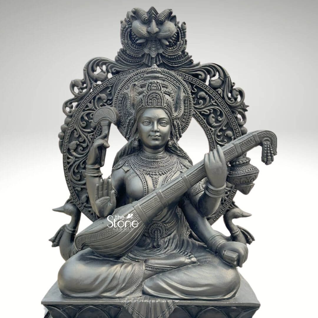 7ft Black Stone Saraswati Statue 2 A magnificent 7-foot black stone Saraswati sculpture is presented from a frontal, slightly elevated perspective against a plain light background. The statue depicts Goddess Saraswati, the deity of knowledge, music, art, speech, wisdom, and learning, seated in a traditional cross-legged posture. She has four arms; her two main hands gracefully play the veena held across her lap. Her upper right hand holds a rosary, and her upper left hand holds a manuscript. The statue is intricately carved from dark black stone, which gives it a powerful and traditional presence. Saraswati is richly adorned with an elaborate crown, intricate necklaces, armlets, earrings, and bangles, all finely detailed. Her garments are sculpted with delicate folds and patterns. Behind her head rises a grand, arching prabhavali featuring exquisite floral and scrollwork carvings, topped with a detailed motif, possibly a Kirtimukha or similar auspicious symbol. On either side of the base of the prabhavali, stylized hamsas her traditional vehicle, are depicted, further emphasizing her iconography. The entire sculpture rests on a multi-tiered rectangular base, also intricately carved. The smooth finish of the black stone highlights the fine artistry and the serene expression of the goddess. This detailed image powerfully conveys the spiritual majesty and artistic craftsmanship of the 7-foot black stone Saraswati statue, making it highly relevant to an article discussing large-scale sculptures for temples or grand spaces.