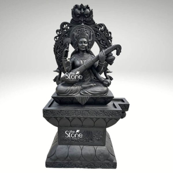 A majestic 7-foot tall Lord Saraswati statue masterfully hand-carved from natural black stone serves as a grand spiritual centerpiece. The Goddess of wisdom, music, and the arts is depicted in a serene seated posture atop a wide lotus pedestal gracefully playing the Veena. Her four arms hold significant sacred symbols: the musical instrument representing harmony, a rosary for meditation, and a book for eternal knowledge. She is framed by an elaborate and massive Prabhavali an ornate stone arch featuring incredibly detailed floral patterns and a protective lion-faced Kirtimukha at the peak. The craftsmanship is evident in the deity's peaceful facial expression, the intricate tiered crown, and the realistic, fluid fabric folds of her traditional attire. This large-scale black stone Saraswati idol with its refined matte charcoal finish, is ideal for grand temple sanctuaries, educational institutions, or luxury home foyers, designed to invite an atmosphere of intellectual clarity and divine grace.