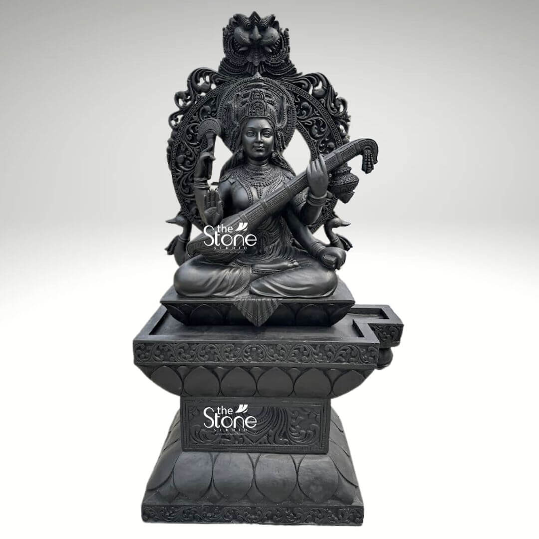 7ft Black Stone Saraswati Statue 1 A majestic 7-foot tall Lord Saraswati statue masterfully hand-carved from natural black stone serves as a grand spiritual centerpiece. The Goddess of wisdom, music, and the arts is depicted in a serene seated posture atop a wide lotus pedestal gracefully playing the Veena. Her four arms hold significant sacred symbols: the musical instrument representing harmony, a rosary for meditation, and a book for eternal knowledge. She is framed by an elaborate and massive Prabhavali an ornate stone arch featuring incredibly detailed floral patterns and a protective lion-faced Kirtimukha at the peak. The craftsmanship is evident in the deity's peaceful facial expression, the intricate tiered crown, and the realistic, fluid fabric folds of her traditional attire. This large-scale black stone Saraswati idol with its refined matte charcoal finish, is ideal for grand temple sanctuaries, educational institutions, or luxury home foyers, designed to invite an atmosphere of intellectual clarity and divine grace.