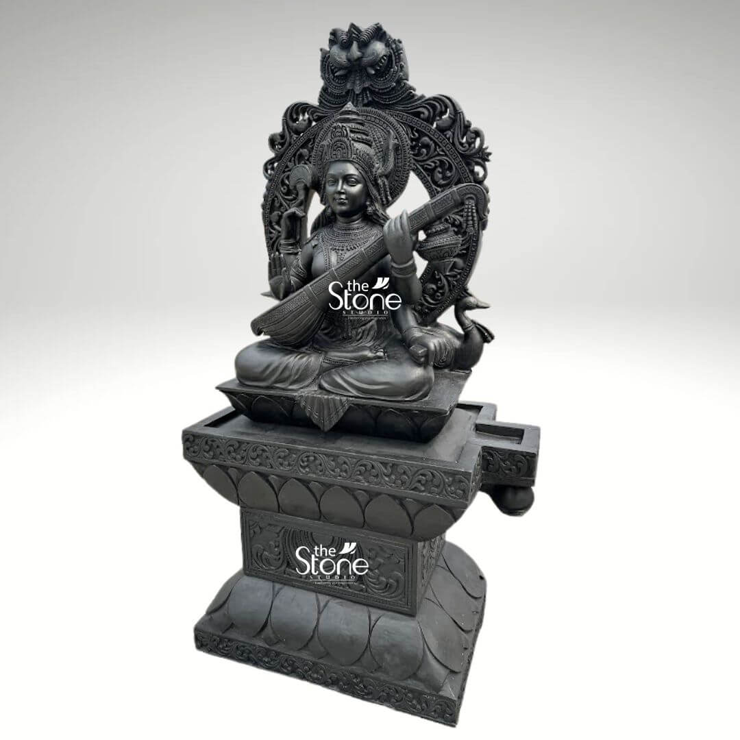 7ft Black Stone Saraswati Statue 4 A majestic 7-foot tall Goddess Saraswati black granite statue masterfully hand-carved from premium natural black stone serving as a grand spiritual centerpiece for a temple foyer or a luxury home entrance. Shown from a three-quarter perspective to highlight its monumental scale and intricate depth, the sculpture depicts the Goddess of knowledge, music, and the arts in a serene seated posture atop an intricate, multi-tiered lotus pedestal. Lord Saraswati is portrayed with four arms, gracefully playing the Veena which symbolizes the harmony of all creative arts, while her upper hands hold a sacred book representing eternal knowledge and a mala for spiritual reflection. She is magnificently framed by a grand, circular Prabhavali featuring deeply carved traditional floral scrolls and a protective Kirtimukha face at its peak. The craftsmanship showcases incredible detail, from the towering tiered crown and large expressive eyes to the layered necklaces, armlets, and realistic fabric folds of her traditional attire. At the base of the statue, her divine vehicle, the swan is delicately rendered in relief. The refined matte charcoal finish of the granite highlights the heritage of traditional Indian stone masonr, making this large-scale Saraswati idol a powerful symbol of wisdom, purity, and creative inspiration designed to invite an aura of intellectual clarity and divine grace into any environment.