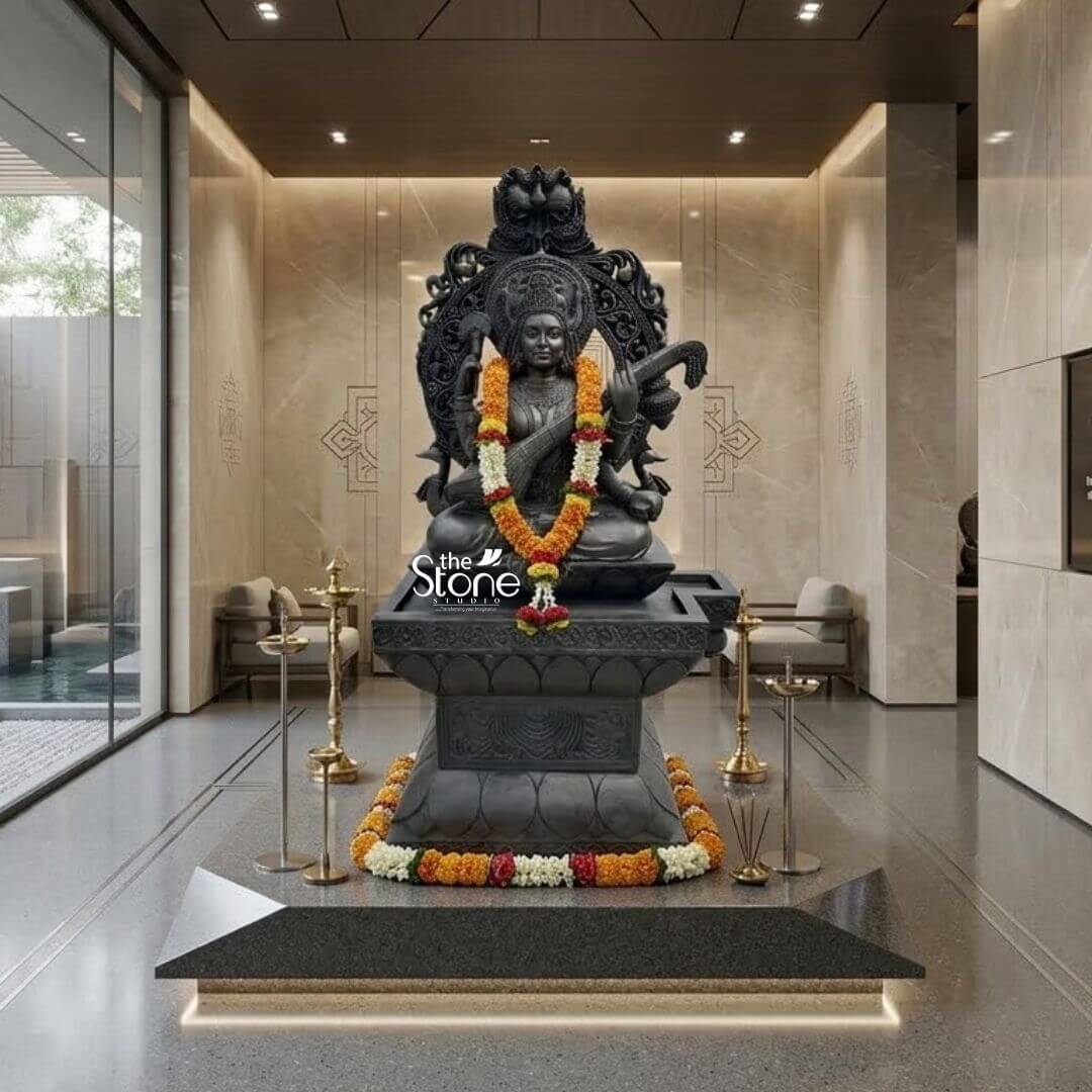 7ft Black Stone Saraswati Statue 3 A majestic 7-foot tall Saraswati Devi statue masterfully hand-carved from natural black stone serves as a grand spiritual centerpiece in a modern, luxury home foyer. The Goddess of knowledge, music, and the arts is depicted in a serene seated posture atop a massive, multi-tiered pedestal adorned with lotus petal relief work. Lord Saraswati is portrayed with four arms, gracefully playing the Veena while her head is framed by an intricately detailed Prabhavali featuring traditional floral motifs and a protective Kirtimukha face at the peak. The craftsmanship showcases incredible detail in her towering tiered crown layered temple jewelry and realistic fabric folds. The statue is elegantly adorned with vibrant orange and yellow marigold garlands. The entire setup is positioned on a polished grey stone floor, flanked by two tall, traditional brass samai oil lamps. The background features minimalist beige marble walls with etched geometric patterns and large floor-to-ceiling glass windows, illustrating a sophisticated fusion of traditional Indian stone craftsmanship and contemporary interior design suitable for a grand temple sanctuary or an upscale residential entrance.
