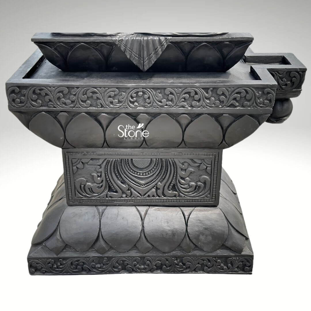 7ft Black Stone Saraswati Statue 5 A robust and intricately carved black stone pedestal, designed as a base for a large statue is presented from a frontal, slightly elevated perspective against a plain light background. The pedestal is constructed in multiple tiers all carved from dark black stone showcasing exquisite traditional craftsmanship. The uppermost section features a wide rectangular platform, with a central stylized lotus design that appears to be a deep oil lamp or offering bowl flanked by smaller indentations for wicks or offerings. The main body of the pedestal is adorned with deeply carved floral and scrollwork patterns along its upper edges, and a repetitive lotus petal motif on its lower sections. The central front face of the lower tier displays a rectangular panel with a highly detailed circular motif, possibly a sun disc or chakra, surrounded by more intricate scrollwork. The entire structure exhibits a textured, matte finish typical of finely carved stone. This detailed image focuses solely on the ornate black stone pedestal, which, in the context of the article, is designed to support a 7-foot Saraswati murti.