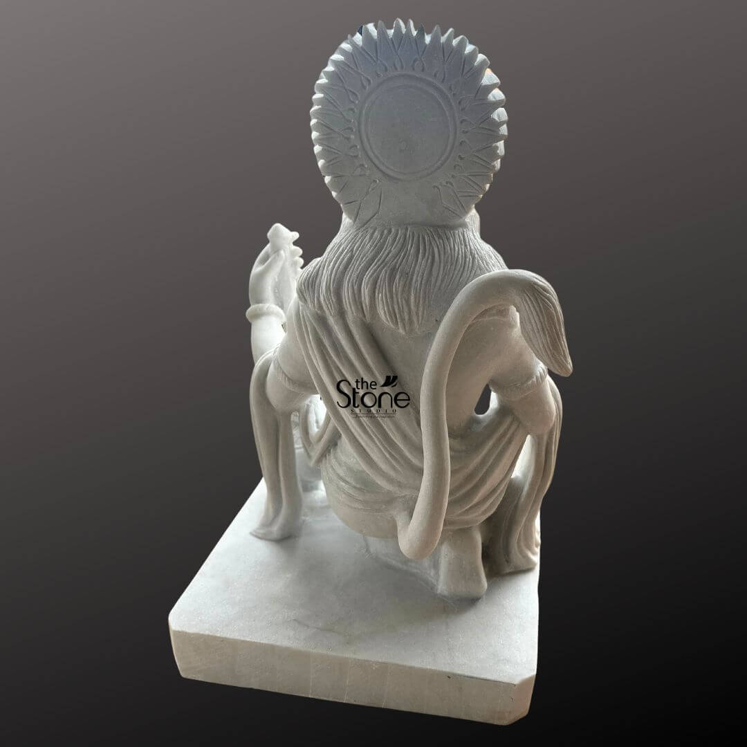 Hanuman Marble Statue for Home Temple 3ft 6 An exquisitely hand-carved 3-foot tall Lord Hanuman marble idol shown from a rear perspective to highlight the exceptional 360-degree craftsmanship of the sculpture. This pure white marble statue depicts the deity in a powerful and devotional kneeling posture making it a majestic spiritual centerpiece for a home puja room. The back view of the sculpture showcases intricate artisan details including the textured flow of Hanuman's hair the realistic, fluid folds of his traditional dhoti, and the iconic, graceful curve of his long tail. Framing the head is a grand, circular Prabhavali with rhythmic sunburst patterns that symbolize divine energy and protection. Crafted from high-quality, polished white marble, the idol rests on a solid rectangular pedestal, ensuring stability and a refined aesthetic. This 36-inch Bajrangbali statue is designed to invite an atmosphere of strength, peace, and unwavering devotion into any sacred prayer space, perfectly blending traditional Vedic iconography with the timeless beauty of natural stone.