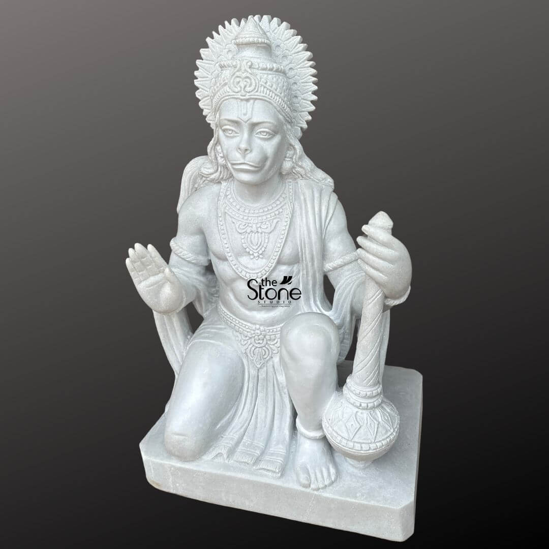 Hanuman Marble Statue for Home Temple 3ft 2 A powerful and intricately carved 3-foot marble Bajrangbali statue is presented from a frontal slightly elevated perspective against a plain dark background. The statue is sculpted from pristine white marble, which highlights every fine detail with remarkable clarity. Lord Hanuman is depicted in a dynamic kneeling posture ready for action yet with a calm and devoted expression. His right hand is raised in a gesture of blessing or fearlessness while his left hand rests on his knee, firmly grasping his gada a symbol of strength and discipline. Hanuman's face is rendered with distinct simian features including expressive eyes and a slightly pointed snout yet conveying profound wisdom and devotion. He is richly adorned with an elaborate crown or headpiece featuring a sun-like halo, intricate necklaces, armlets, anklets, and a sacred thread across his chest. His muscular physique is subtly sculpted covered by finely draped garments that show detailed folds and patterns. The entire statue rests on a simple, rectangular marble base. The smooth, lustrous finish of the white marble emphasizes the exquisite artistry and the powerful, benevolent presence of Lord Hanuman. This detailed image vividly conveys the spiritual majesty and artistic craftsmanship of the marble Bajrangbali statue, making it highly relevant to an article discussing such revered idols.