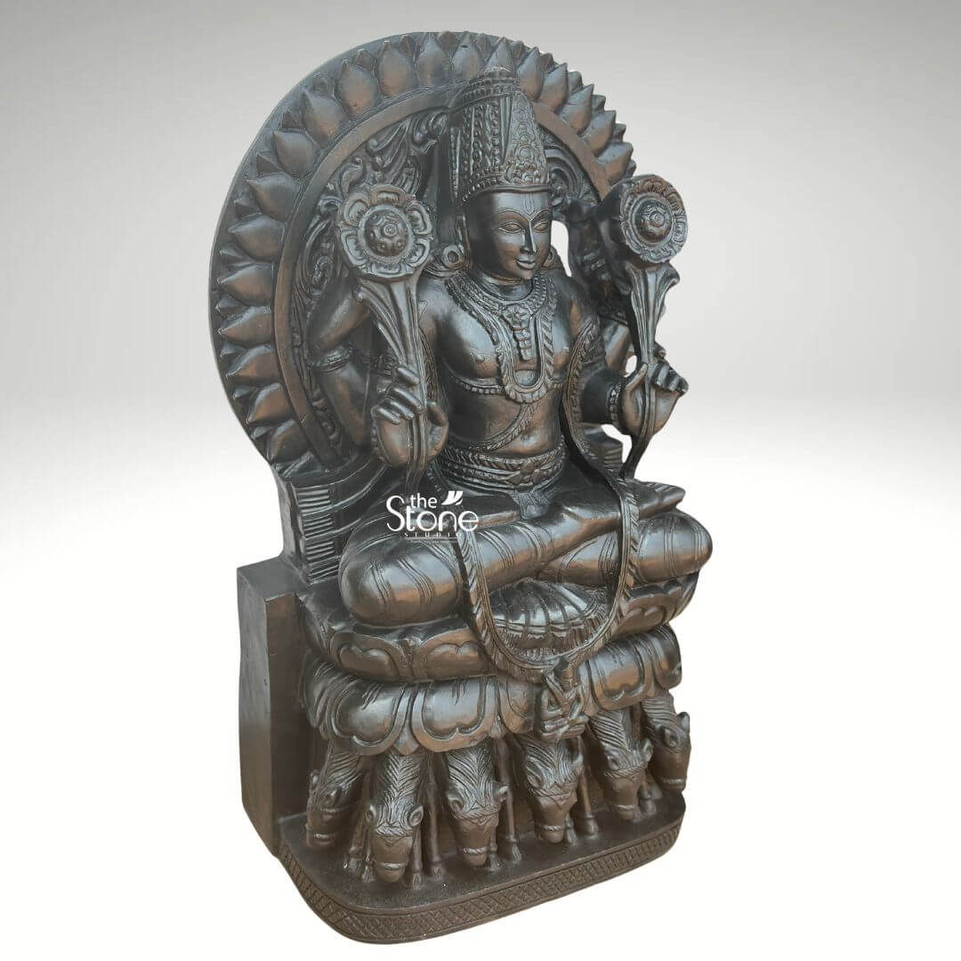 Surya Dev Black Stone Statue 30" 2 An exquisitely hand-carved 2.5-foot tall Lord Surya Dev idol masterfully sculpted from premium natural black stone with a refined matte charcoal finish. This majestic spiritual masterpiece depicts the Sun God in a traditional seated posture atop an intricate multi-tiered lotus pedestal. Lord Surya Dev is portrayed with two arms holding sacred blooming lotus blossoms symbolizing divinity and creation. He is adorned with a towering tiered crown mukut layered necklaces and armlets that showcase the exceptional craftsmanship of traditional Indian stone masonry. The statue features a grand, circular Prabhavali embellished with rhythmic petal-like relief patterns that symbolize a divine sunburst aura. At the base of the sculpture, the seven horses of his celestial chariotSaptashva are meticulously carved in high relief signifying the seven days of the week and the seven colors of light. This 30-inch black stone Surya Dev statue serves as a profound spiritual icon, ideal for a home mandir foyer, or meditation space to invite energy, clarity, and positive energy into the environment.