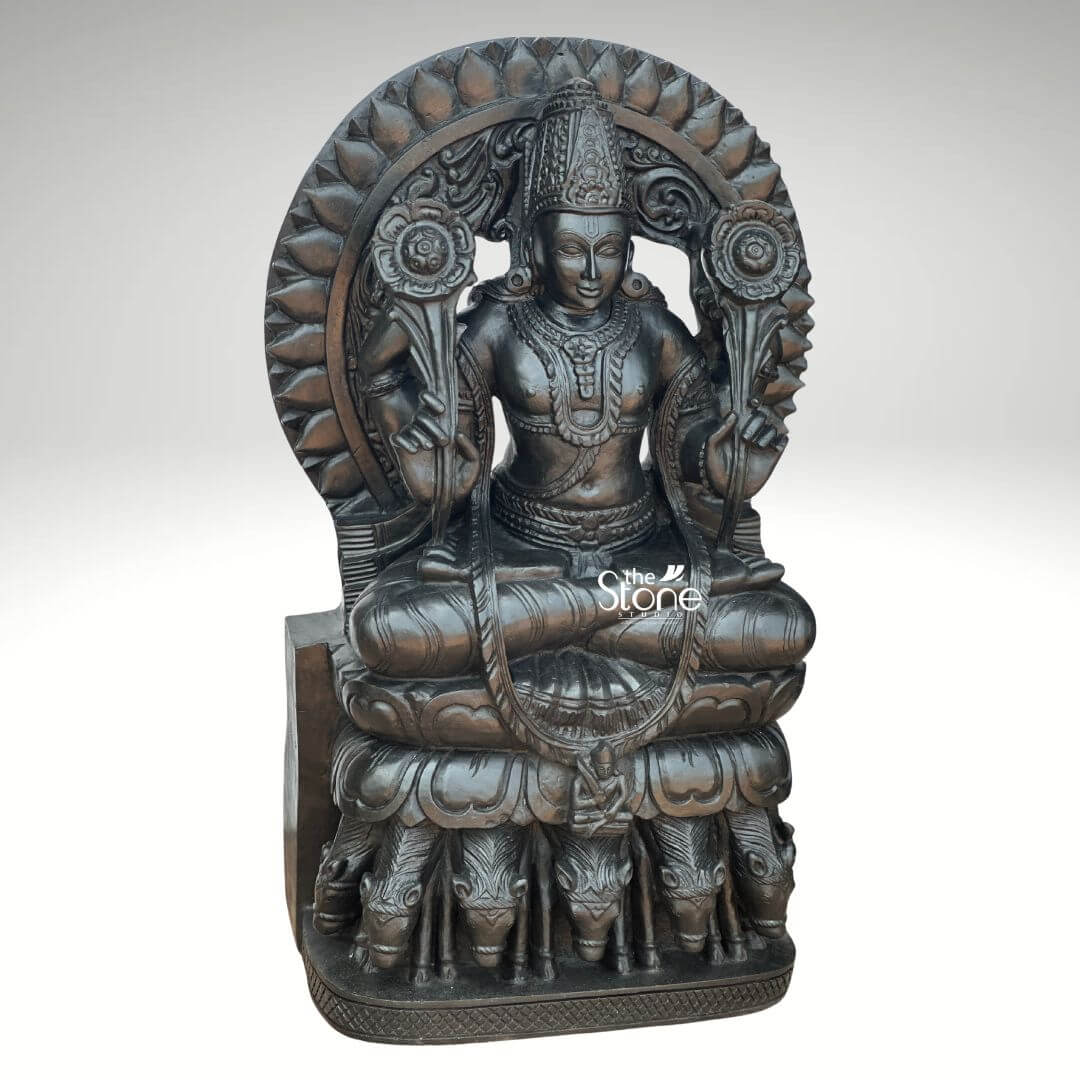 Surya Dev Black Stone Statue 30" 4 A majestic 2.5-feet black stone Surya Dev statue is presented from a frontal slightly elevated perspective against a plain light background. The sculpture depicts Surya Dev the Hindu solar deity, seated in a regal posture embodying power and enlightenment. The entire statue is meticulously carved from rich, dark black stone, which gives it a profound and traditional presence with a subtle antique-like sheen. Surya Dev is shown with two arms each holding a blooming lotus flower symbols of purity and creation extending upwards from his shoulders. He is richly adorned with an elaborate crown intricate necklaces armlets, earrings and a sacred thread across his chest. His serene face with slightly downcast eyes, radiates benevolence. Behind his head rises a grand, circular prabhavali intricately carved with lotus petals radiating outwards symbolizing the sun's rays and his divine effulgence. The most distinctive feature of the statue is its base: Surya Dev is seated atop a chariot driven by seven horses, which are exquisitely carved in a row beneath his lotus throne representing the seven colors of the rainbow or the seven days of the week. The smooth yet detailed finish of the black stone highlights the fine artistry and the powerful, benevolent presence of the solar deity. This detailed image powerfully conveys the spiritual majesty and artistic craftsmanship of the black stone Surya Dev statue, making it highly relevant to an article discussing such revered idols.