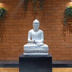 A serene 3-foot tall Lord Buddha stone statue masterfully hand-carved from natural white sandstone serves as a spiritual centerpiece in a Raichur residence. The sculpture depicts Buddha in a peaceful meditative posture seated in the traditional lotus position atop an intricately detailed multi-tiered lotus pedestal. The deity’s hands are held in the Dhyana Mudra the universal gesture of meditation symbolizing deep concentration and inner peace. The statue is elegantly positioned on a solid black granite platform creating a striking contrast against a rustic reddish-brown brick wall backdrop. Small, lush green hanging plants on either side of the wall add a touch of nature and life to the scene. This setting perfectly illustrates how a big-sized stone Buddha idol can be integrated into residential or commercial landscapes in Raichur Karnataka, to create a tranquil Zen sanctuary for mindfulness and reflection. The smooth texture and pure white finish of the sandstone Buddha radiate a sense of divine grace and timeless elegance.