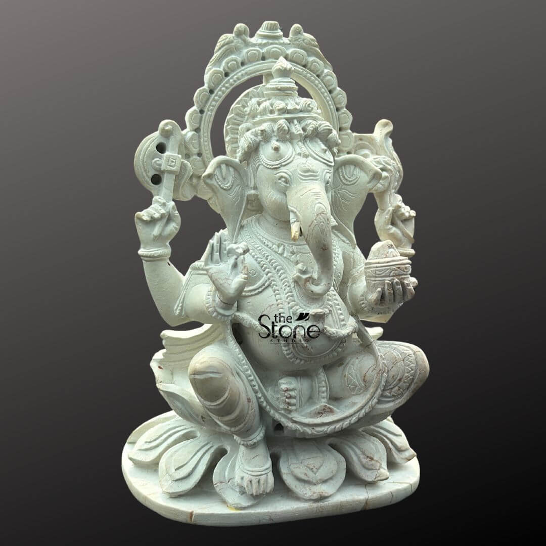 Small Ganesha Idol for Car Dashboard 4" 2 A beautifully detailed white stone Ganesha statue, suitable for a car dashboard, is presented against a plain dark background. The statue depicts Lord Ganesha in a seated, somewhat squatting posture on a lotus base, radiating an aura of wisdom and benevolence. He has four arms; his upper right hand holds an axe or goad, while his upper left hand grasps a noose (not fully visible). His lower right hand is raised in a gesture of blessing (Abhaya Mudra), and his lower left hand holds a small bowl of modaks (sweet dumplings), with his trunk curving towards it, indicating his fondness for sweets. The white stone material highlights the intricate carvings, including the elaborate crown, jewelry, and draped dhoti. A decorative halo or prabhavali with a serrated edge frames Ganesha's head, further enhancing his divine presence. The texture of the stone is smooth, emphasizing the fine artistry. This detailed image clearly illustrates the sacred and decorative qualities of the white stone Ganesha statue, making it ideal for an article discussing dashboard placement.