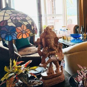 An exquisitely hand-carved 15-inch pinkstone statue of Dancing Ganesha known as Nritya Ganesha serves as a majestic spiritual centerpiece in a contemporary Kolkata home. The idol is masterfully sculpted from natural pink stone showcasing a warm, earthy tone and intricate artisan details in the deity's tiered crown, traditional jewelry, and flowing garments. Lord Ganesha is depicted in an energetic dance pose with one leg raised in a rhythmic stance and his trunk curved gracefully toward his hand, standing atop an ornate oval lotus pedestal. The sculpture is artfully placed on a dark wooden surface in a well-lit living area, positioned next to a large, vibrant Tiffany-style lamp featuring a colorful floral glass shade. Flanked by lush indoor foliage in the foreground, the Dancing Ganesha statue is set against a background of a sun-drenched balcony and sophisticated interior decor. This arrangement perfectly illustrates how a traditional pinkstone Ganesha idol can be integrated into modern Kolkata interior design to invite joy prosperity and a sense of sacred heritage into an urban living space.